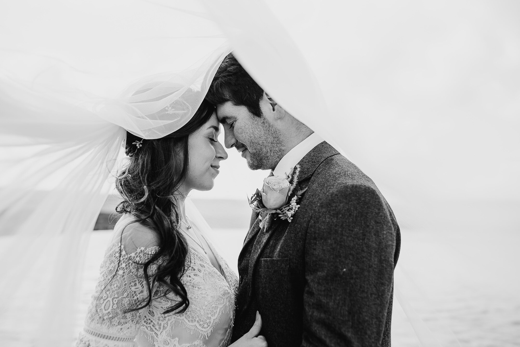 Bride and groom embracing under a veil in black and white wedding photo.