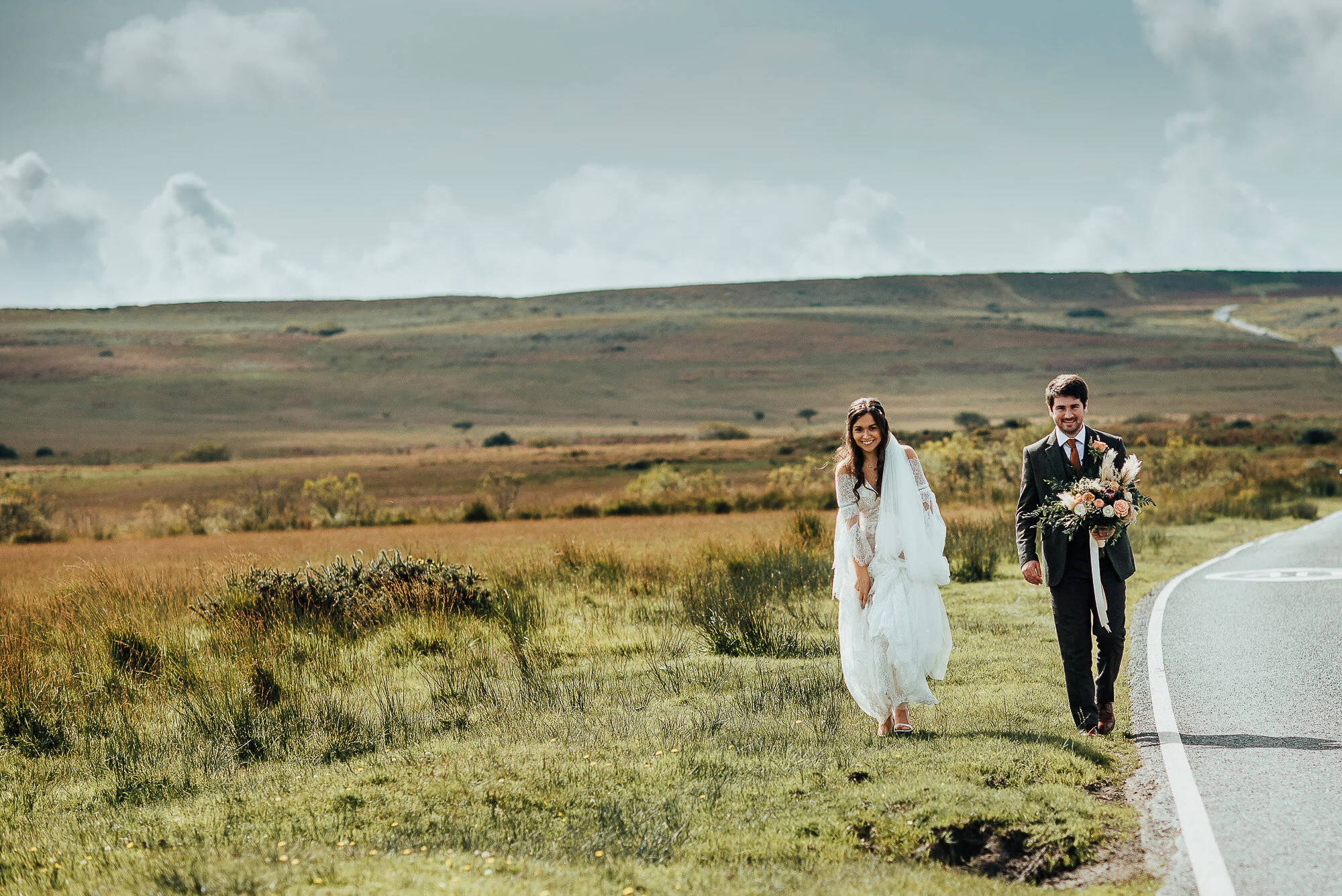 Bride and groom walking on a rural road with a scenic landscape and cloudy sky in the background.