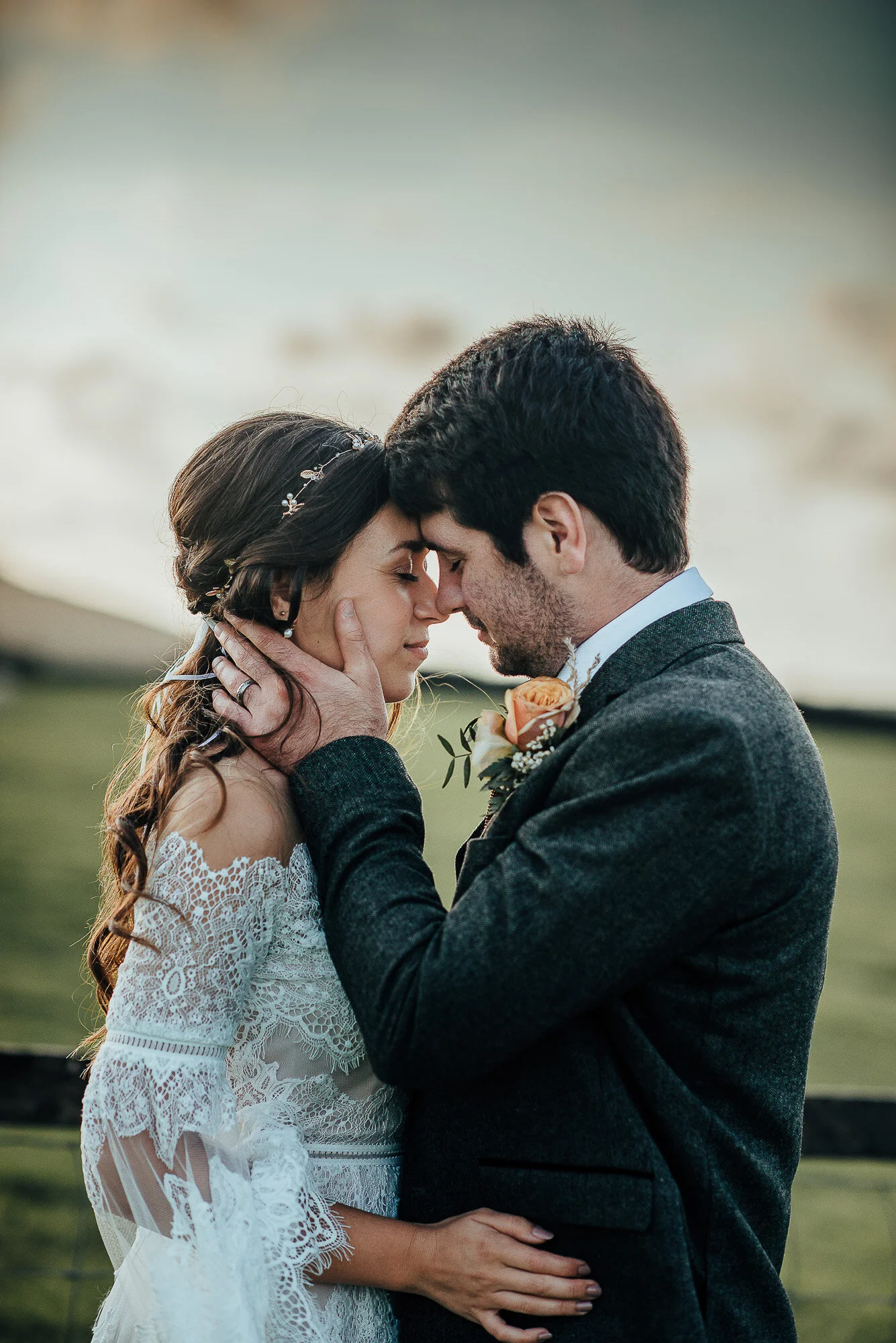 Romantic couple embracing outdoors; woman in lace dress, man in suit with boutonniere.