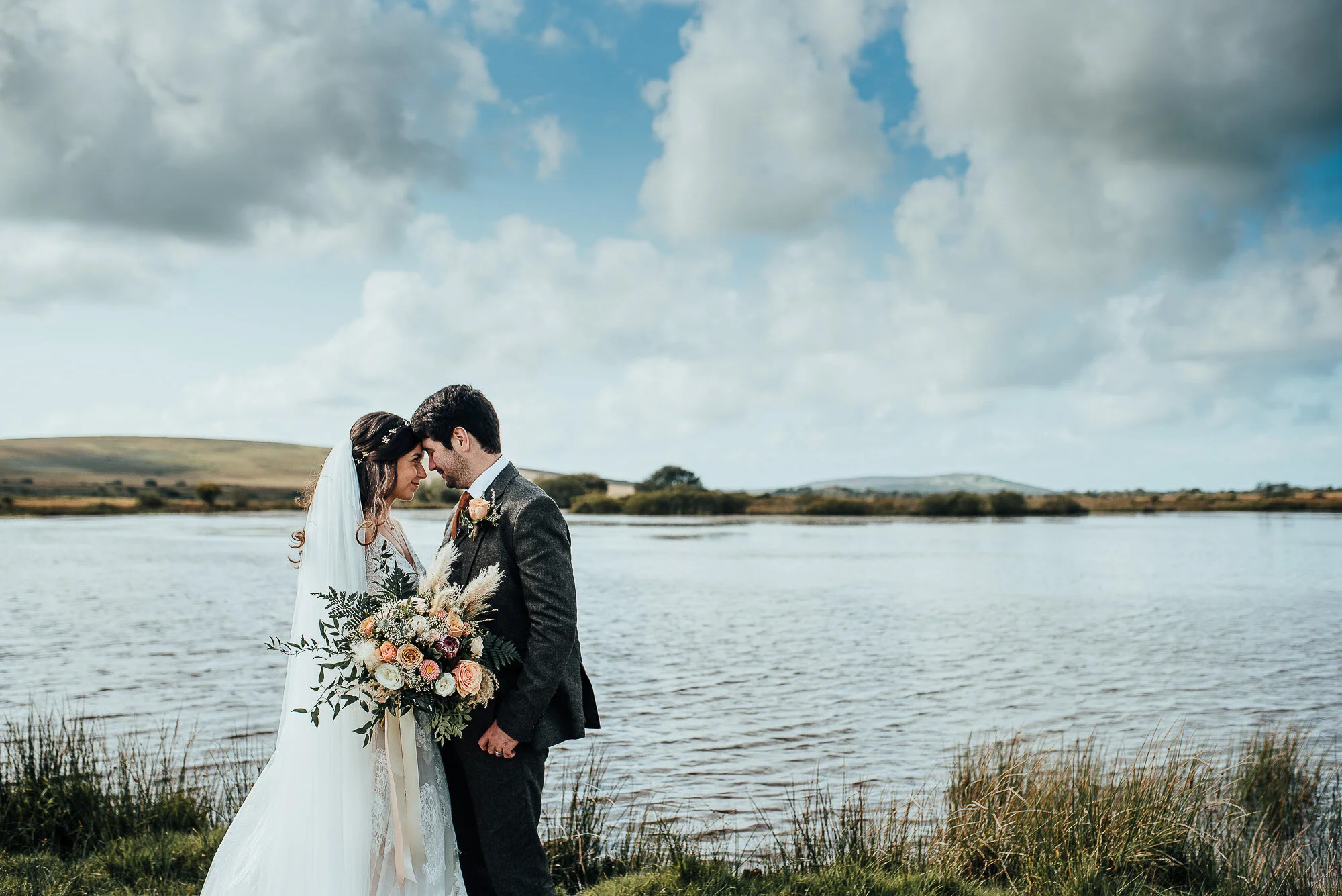 Bride and groom standing by a lake with clouds in the sky