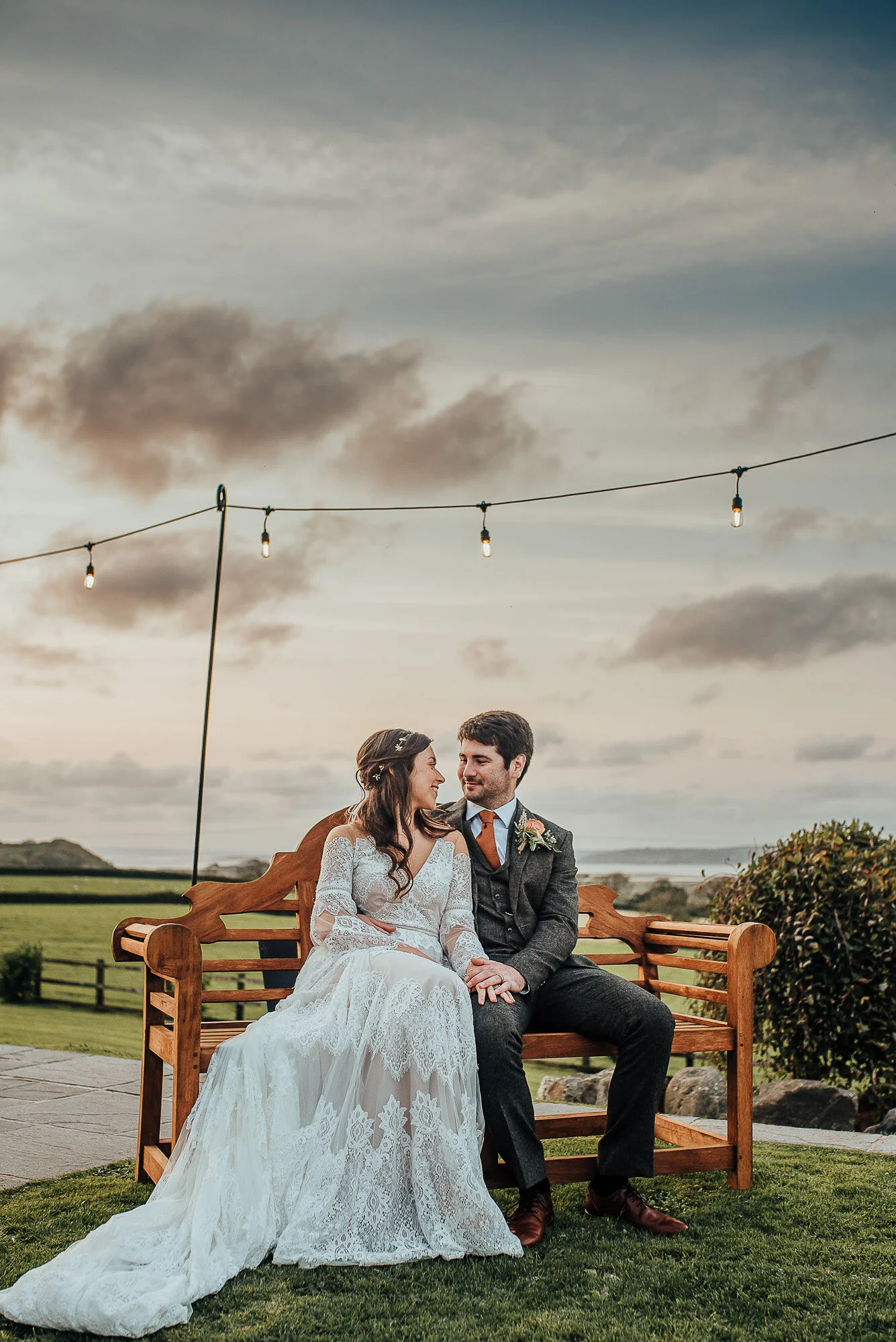 Bride and groom sitting on a wooden bench outdoors, with string lights above them, surrounded by a scenic landscape and a cloudy sky.