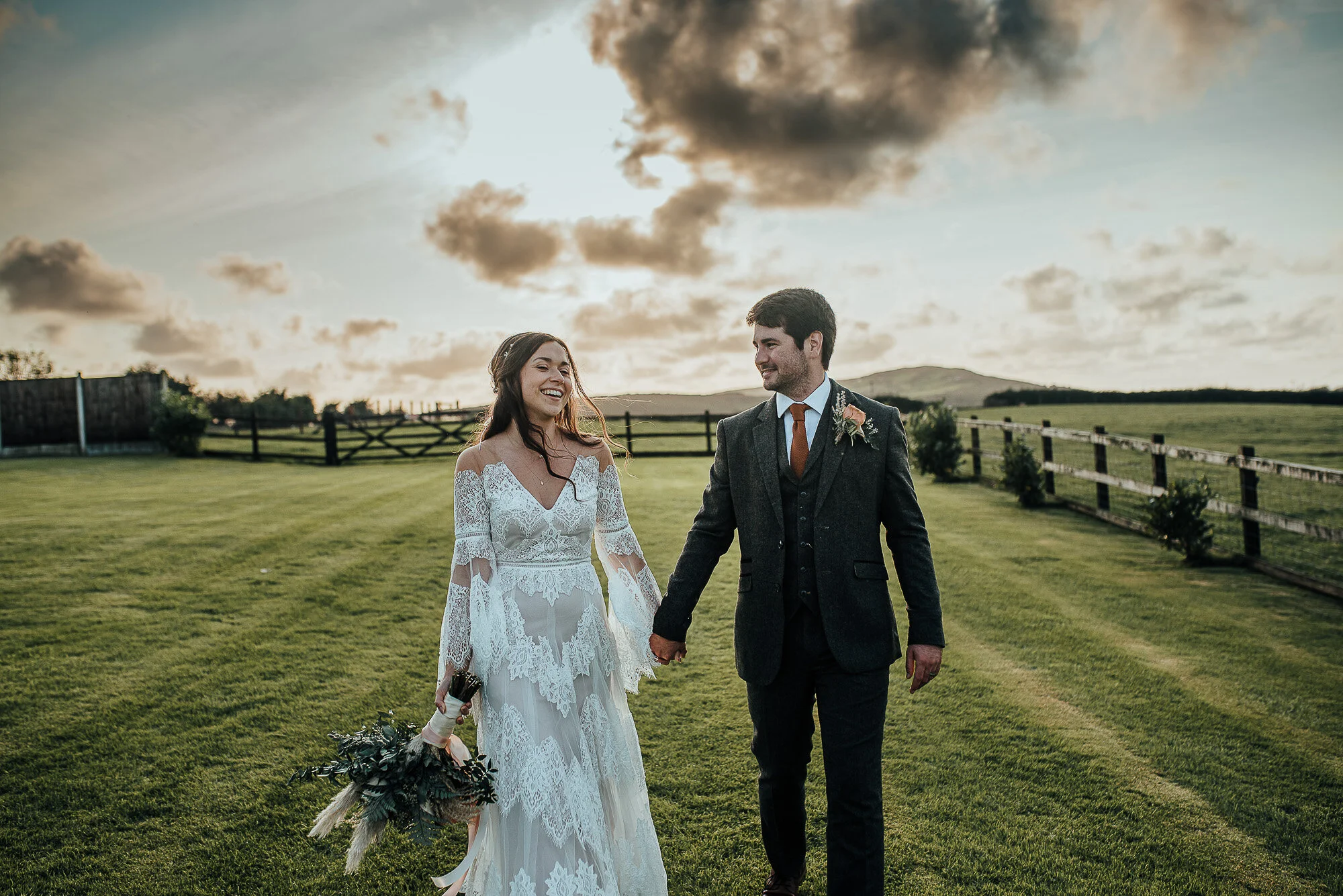 Bride and groom walking hand-in-hand on a grassy field during sunset, with a fence and hills in the background.