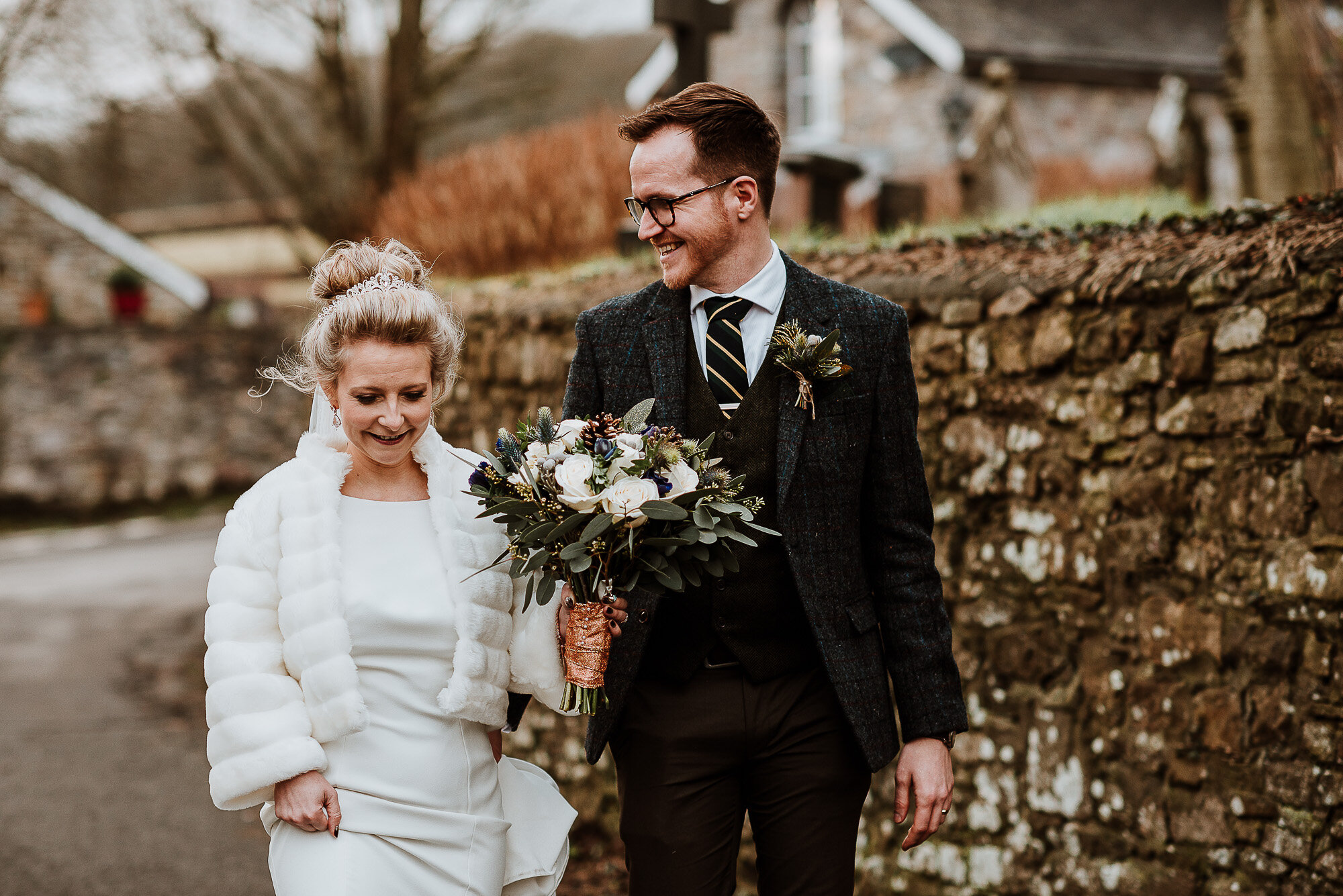 Bride and groom walking together, bride in a white dress and fur wrap, groom in a suit, holding a bouquet, with a stone wall and rural background.