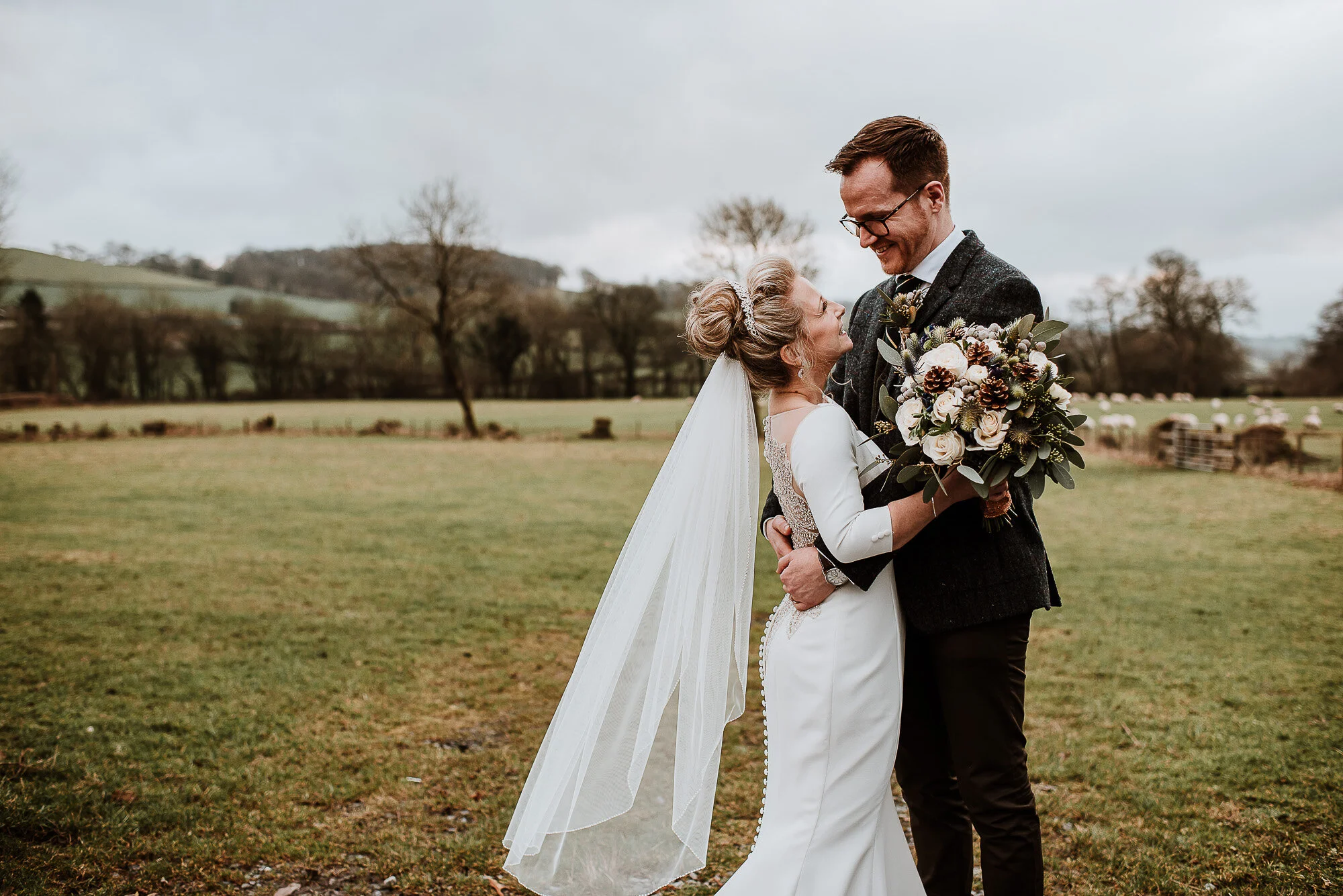 Bride and groom embracing in a field with a scenic landscape background, the bride holding a bouquet and wearing a veil.