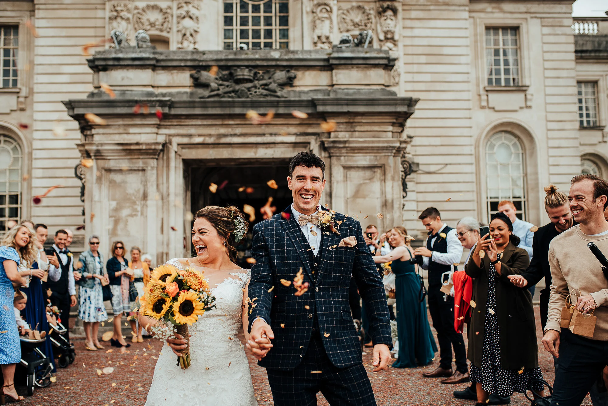 A newlywed couple smiling and holding hands while walking through a crowd of celebrating guests outside a historic building. The bride is holding a sunflower bouquet, and confetti is being thrown. People around are clapping and taking photos.