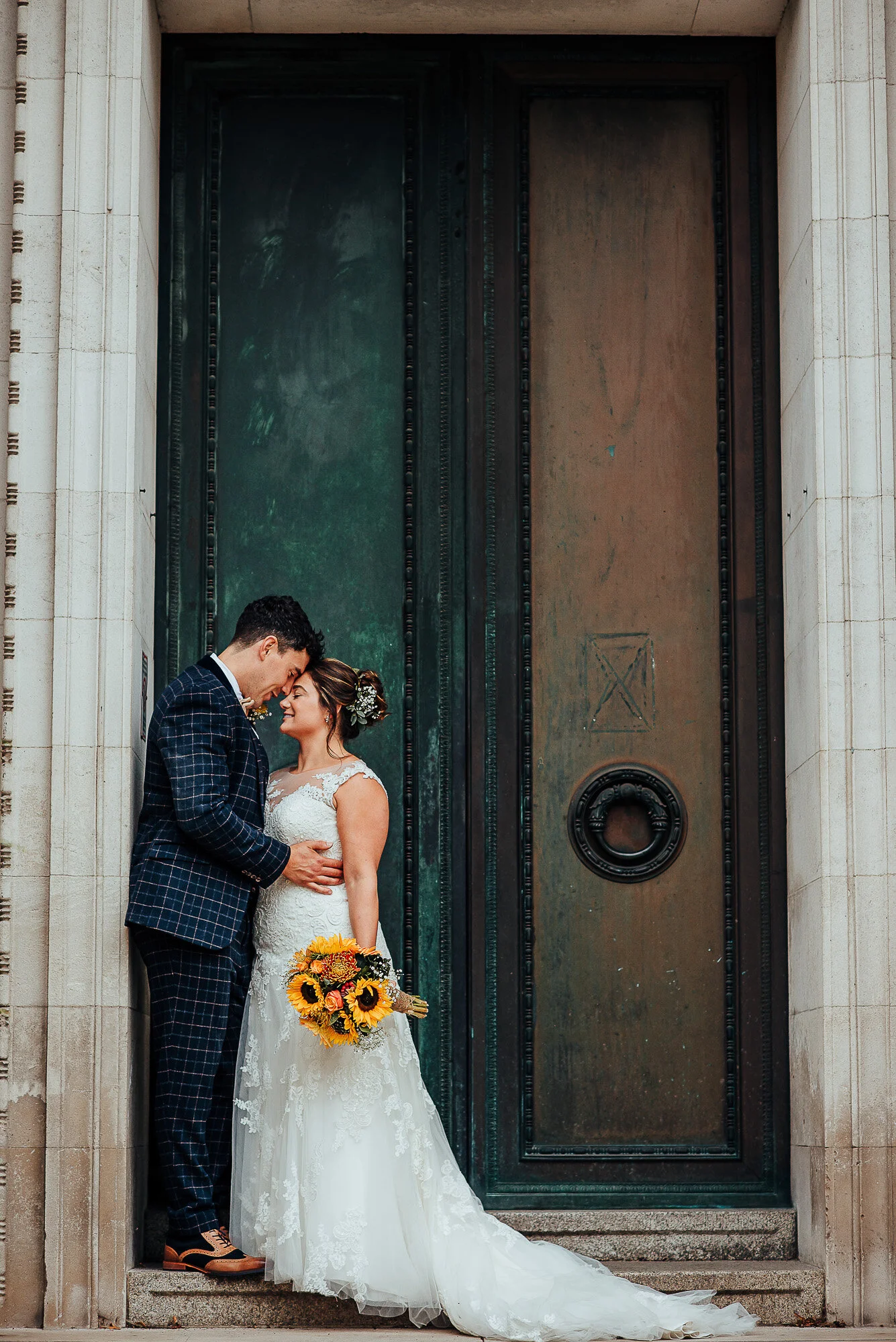 Bride and groom embracing in front of large green door, bride holding sunflower bouquet, groom in checkered suit, bride in lace dress.
