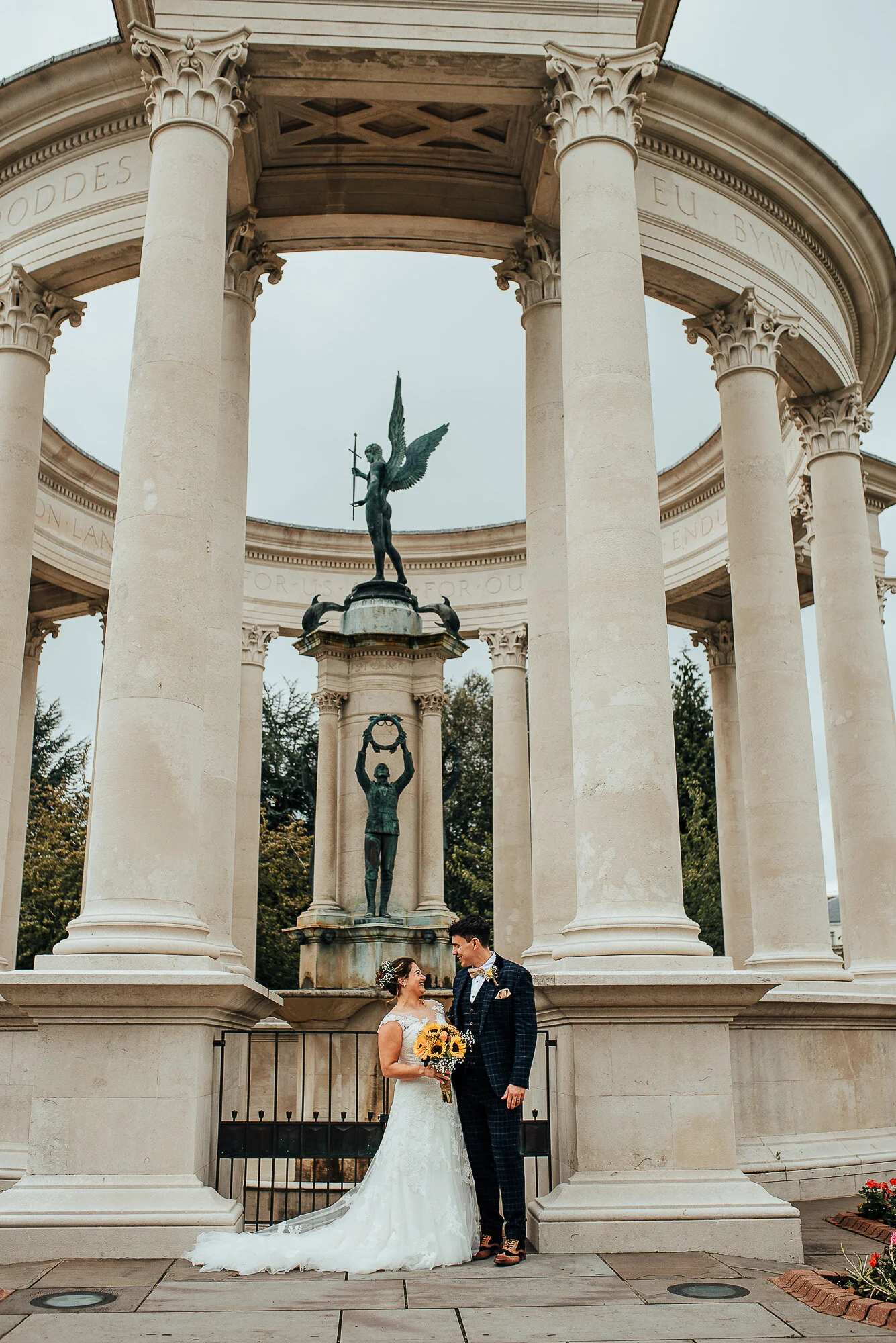 Bride and groom under a neoclassical monument with tall columns, featuring a winged statue on top. The bride holds a bouquet of sunflowers.