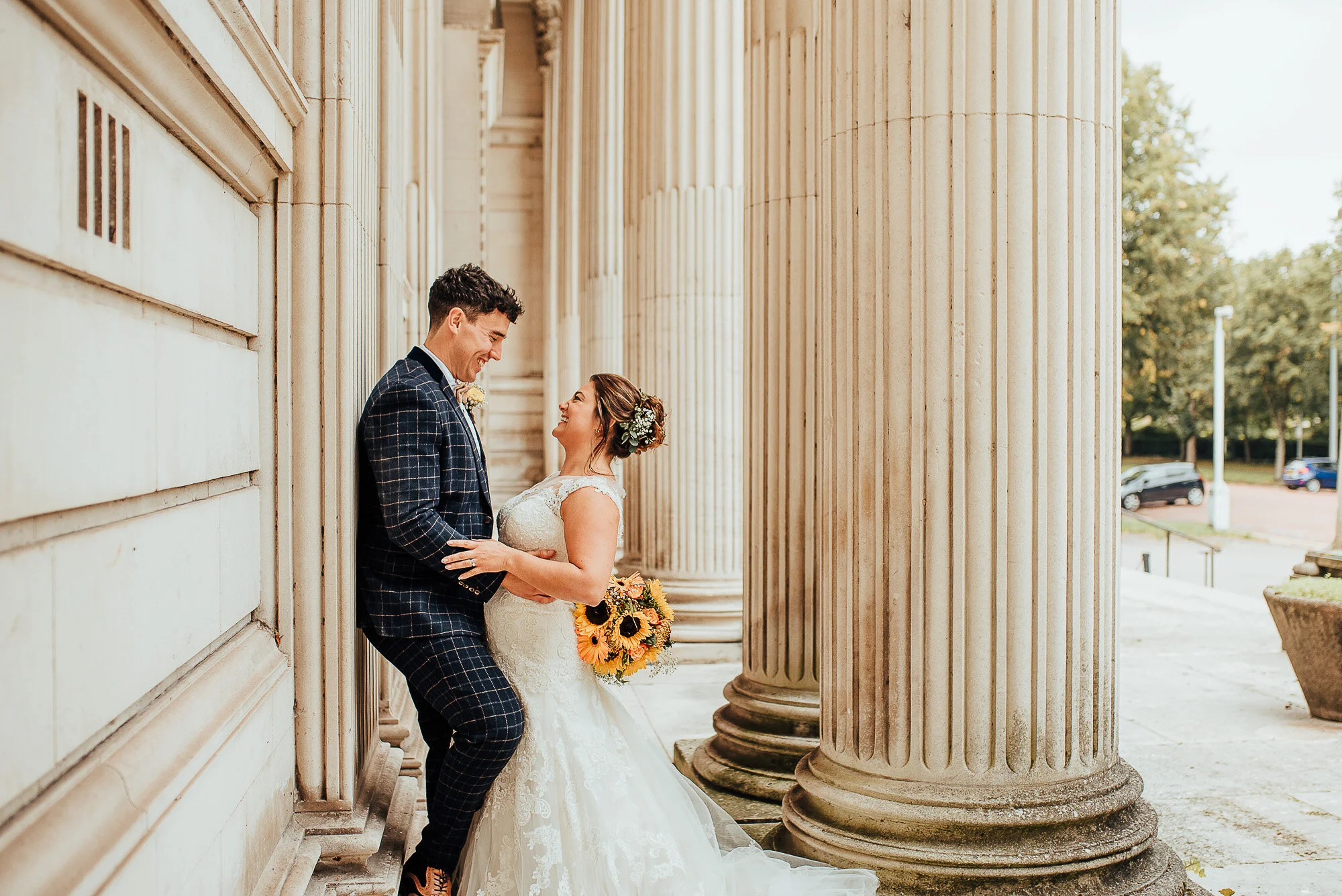 Bride and groom laughing and embracing in front of stone columns, bride holding sunflower bouquet.