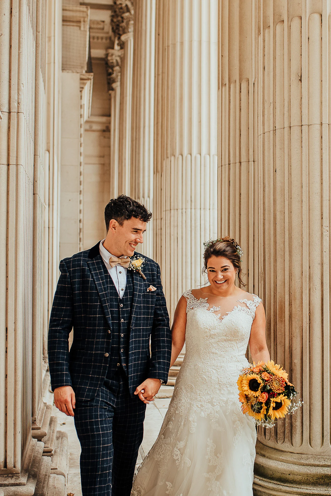 Bride and groom smiling while walking between tall columns, bride holding a bouquet of sunflowers.