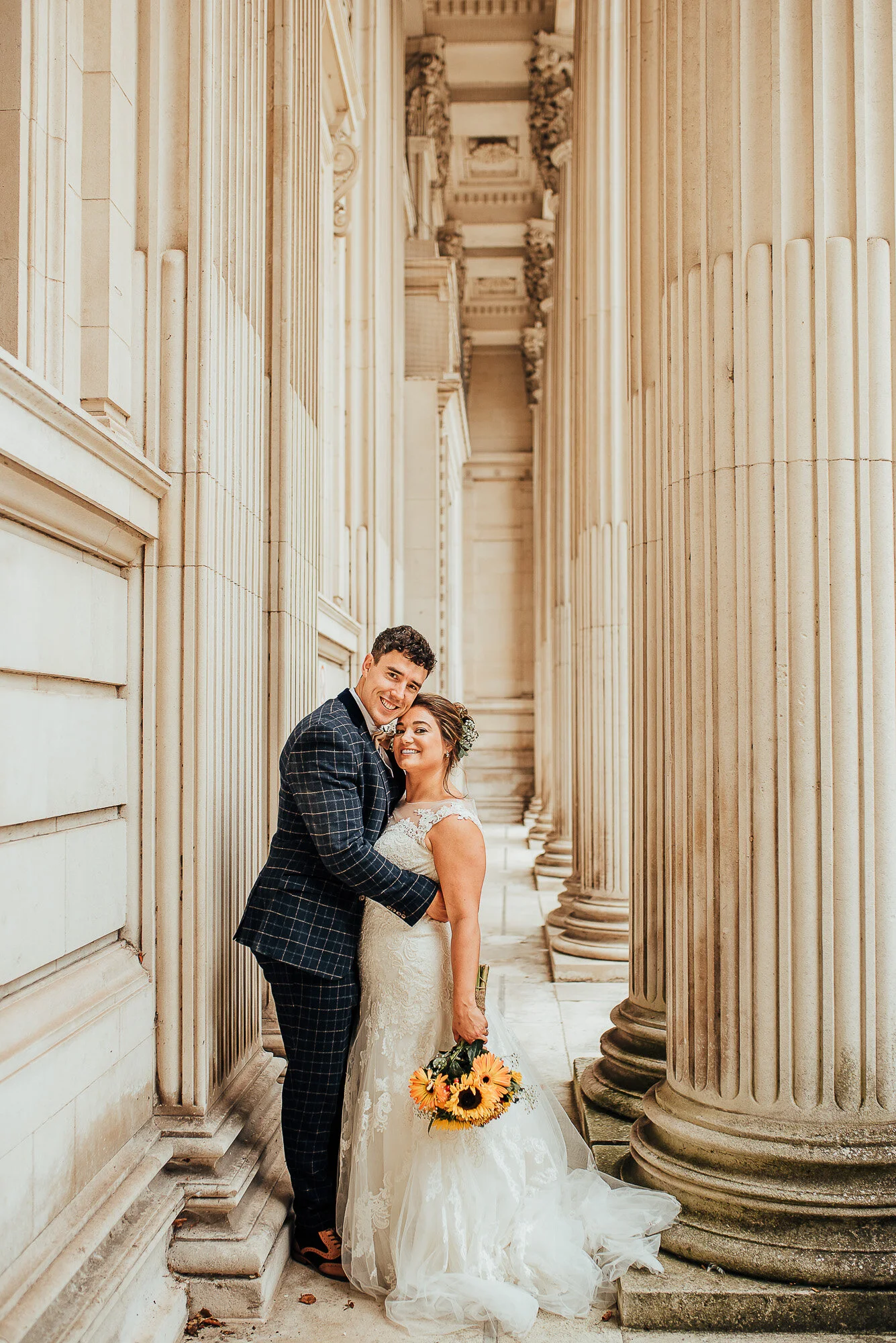 A smiling couple embracing in formal attire, with the bride holding a bouquet of sunflowers, standing in a classical column-lined corridor.