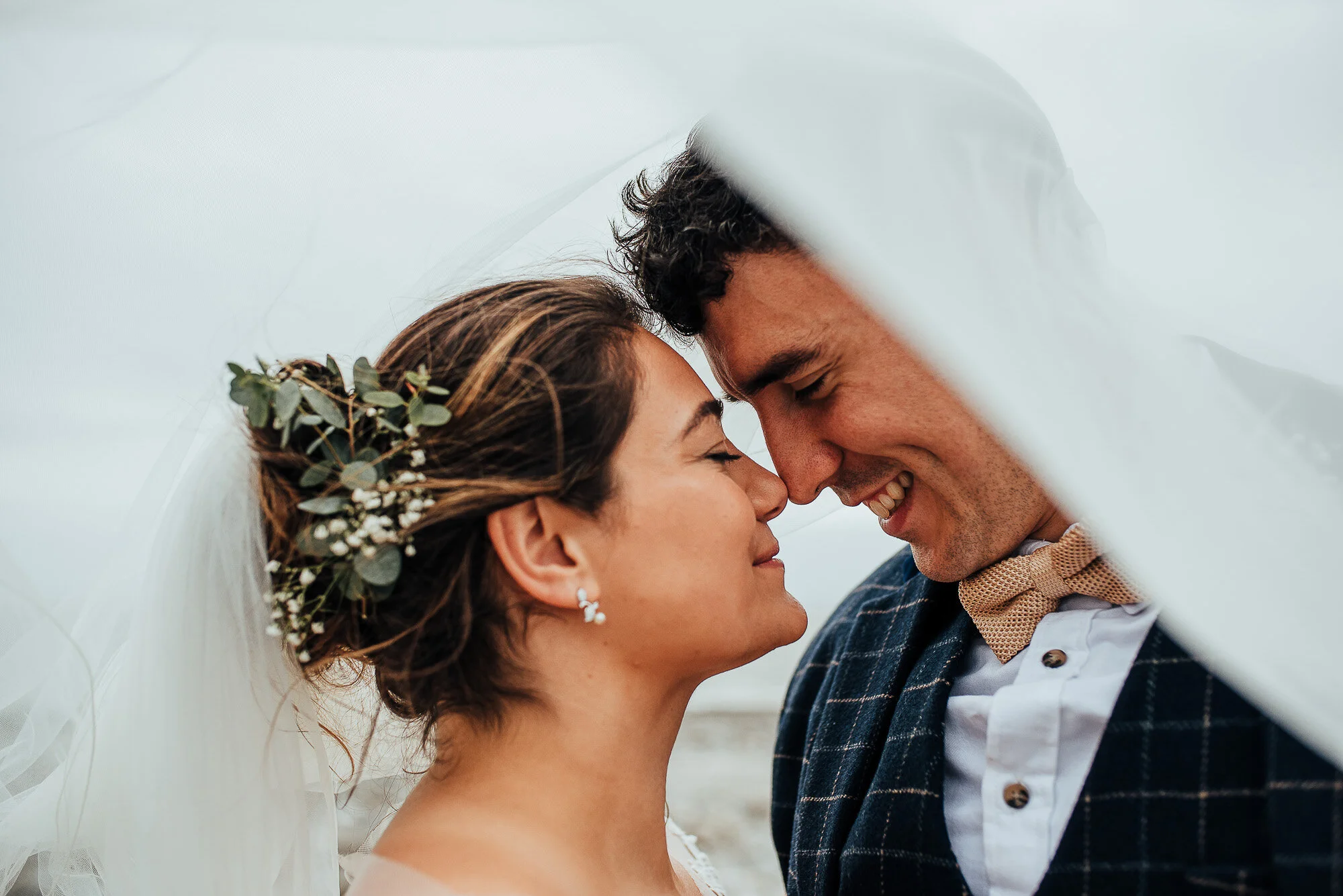 Bride and groom smiling under a veil, with greenery in the bride's hair.