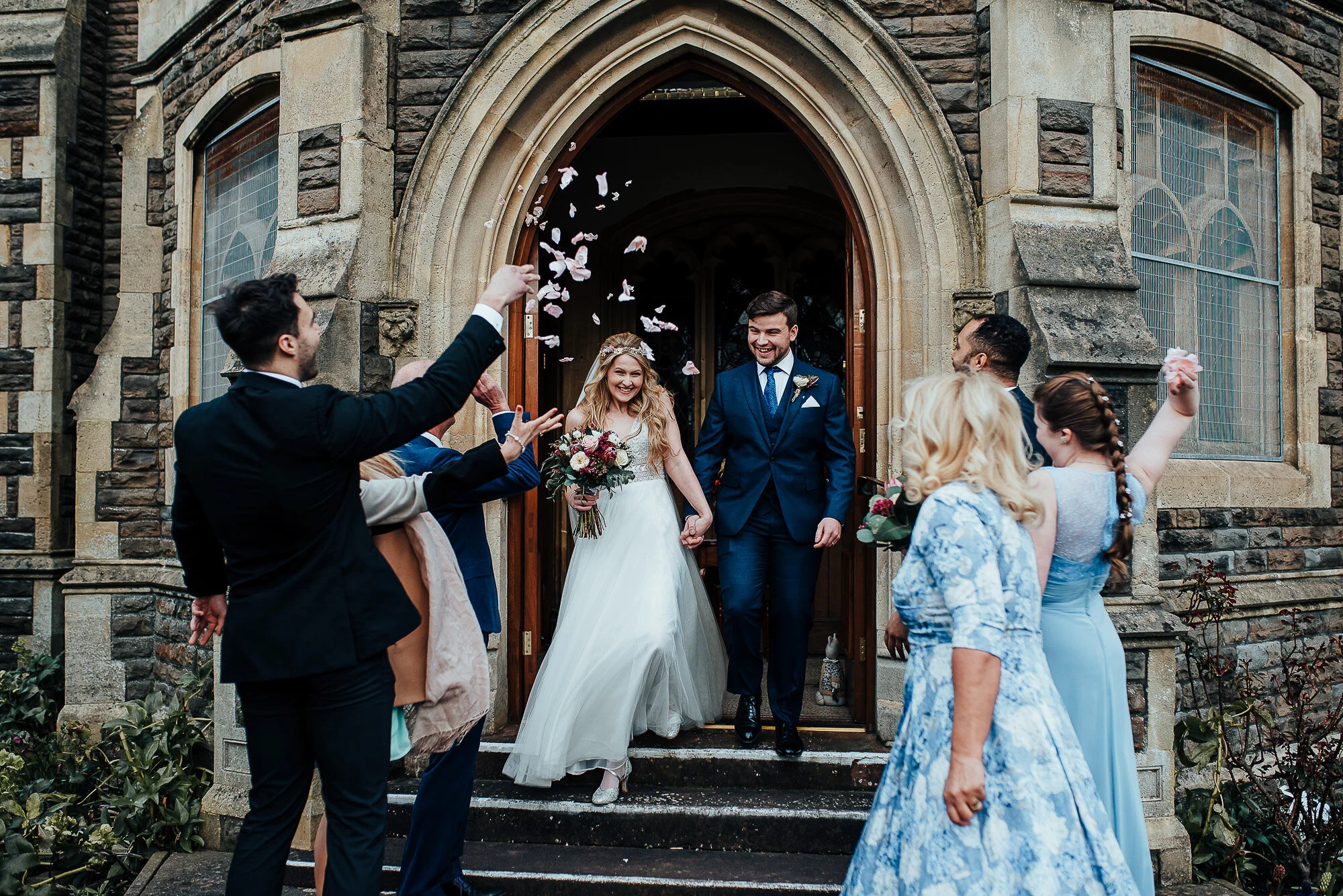 Newlywed couple exiting church, guests throwing flower petals, groom in blue suit, bride in white dress with bouquet, surrounded by smiling guests in formal attire.