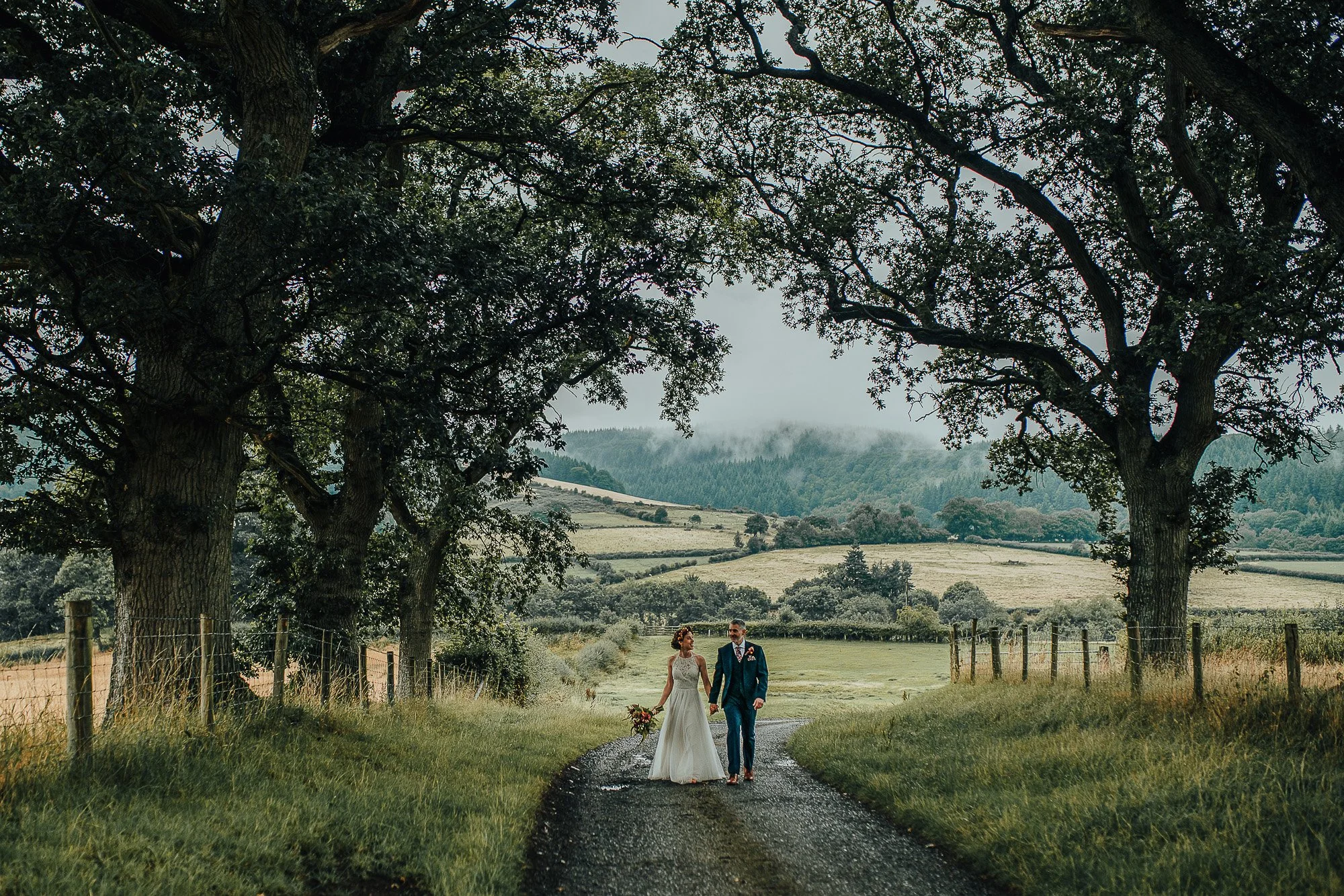 Bride and groom walking on a country road surrounded by trees and fields