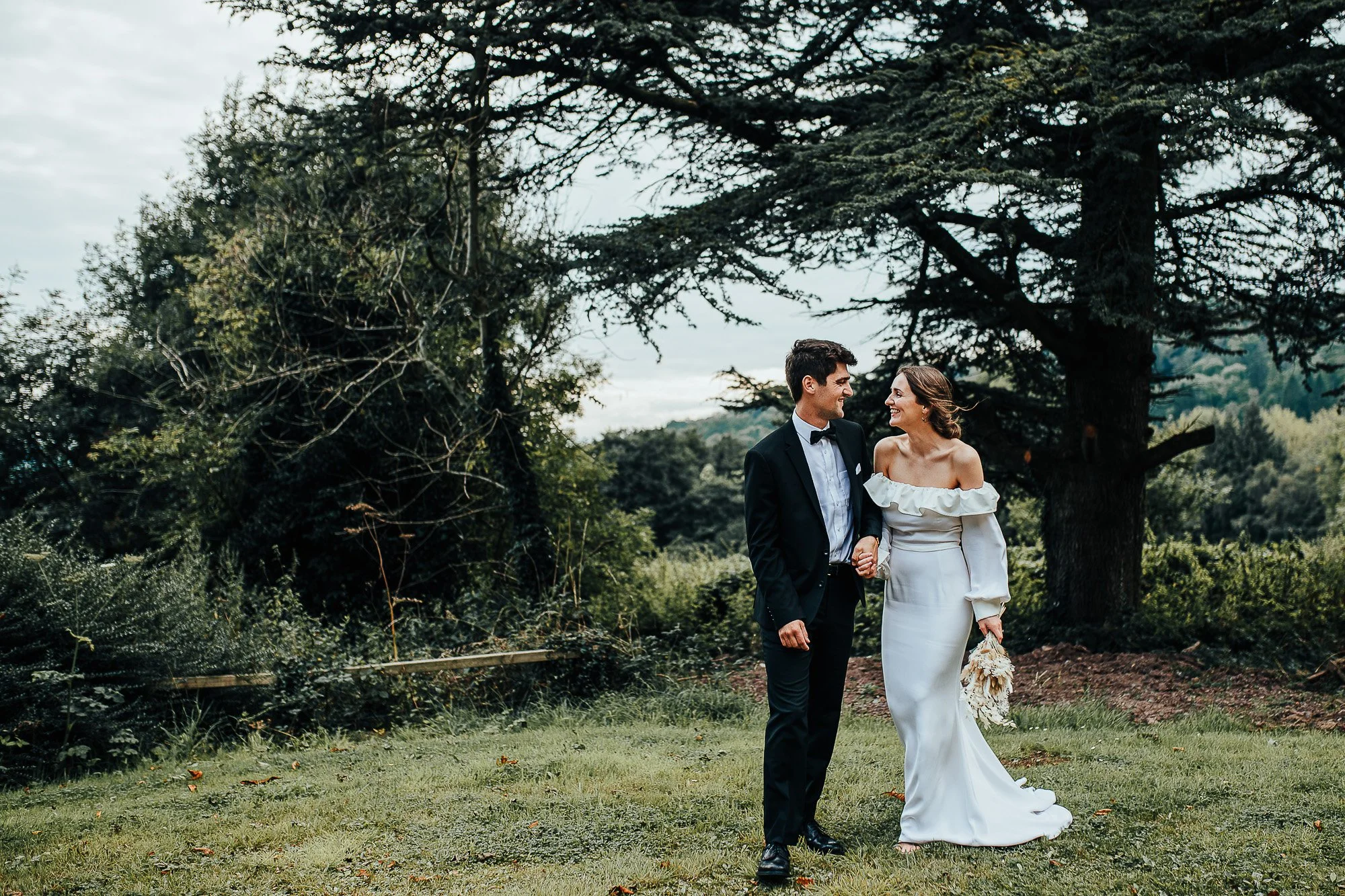 Bride and groom in a wooded outdoor setting, smiling and holding hands; bride in white dress, groom in dark suit.