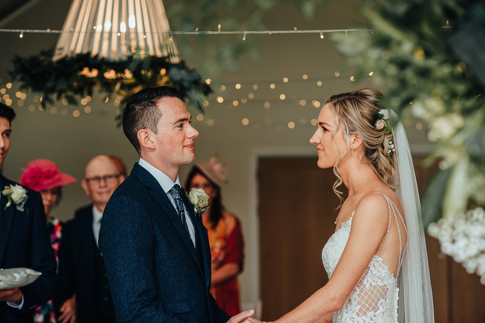 A couple getting married, exchanging vows indoors. The groom is in a formal suit, and the bride is in a lace wedding gown with a veil. They are holding hands, surrounded by decorative lighting and greenery, with guests in the background.