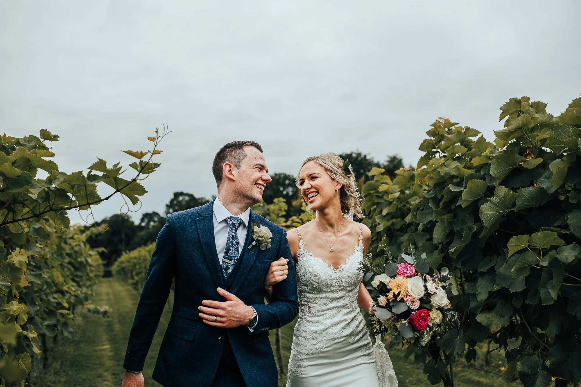 Bride and groom walking through a vineyard, the bride holding a bouquet of flowers, both smiling.