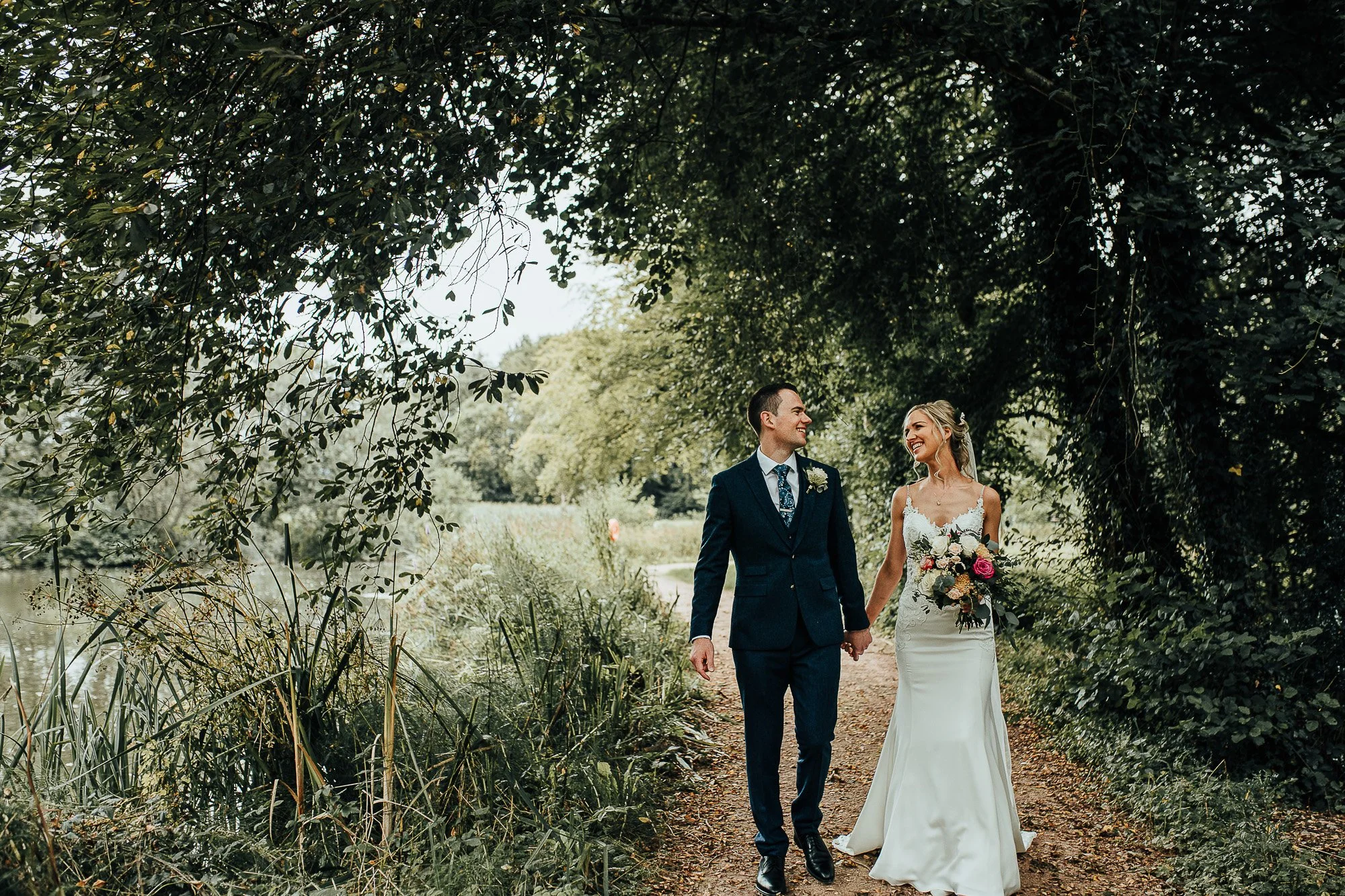 Bride and groom walking hand in hand on a wooded path, with bride holding a bouquet and smiling at each other.