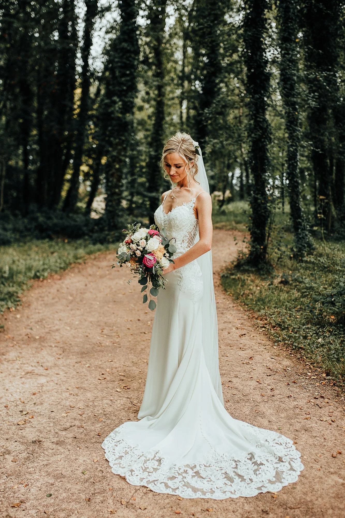 Bride in white wedding dress holding bouquet, standing on a forest path.