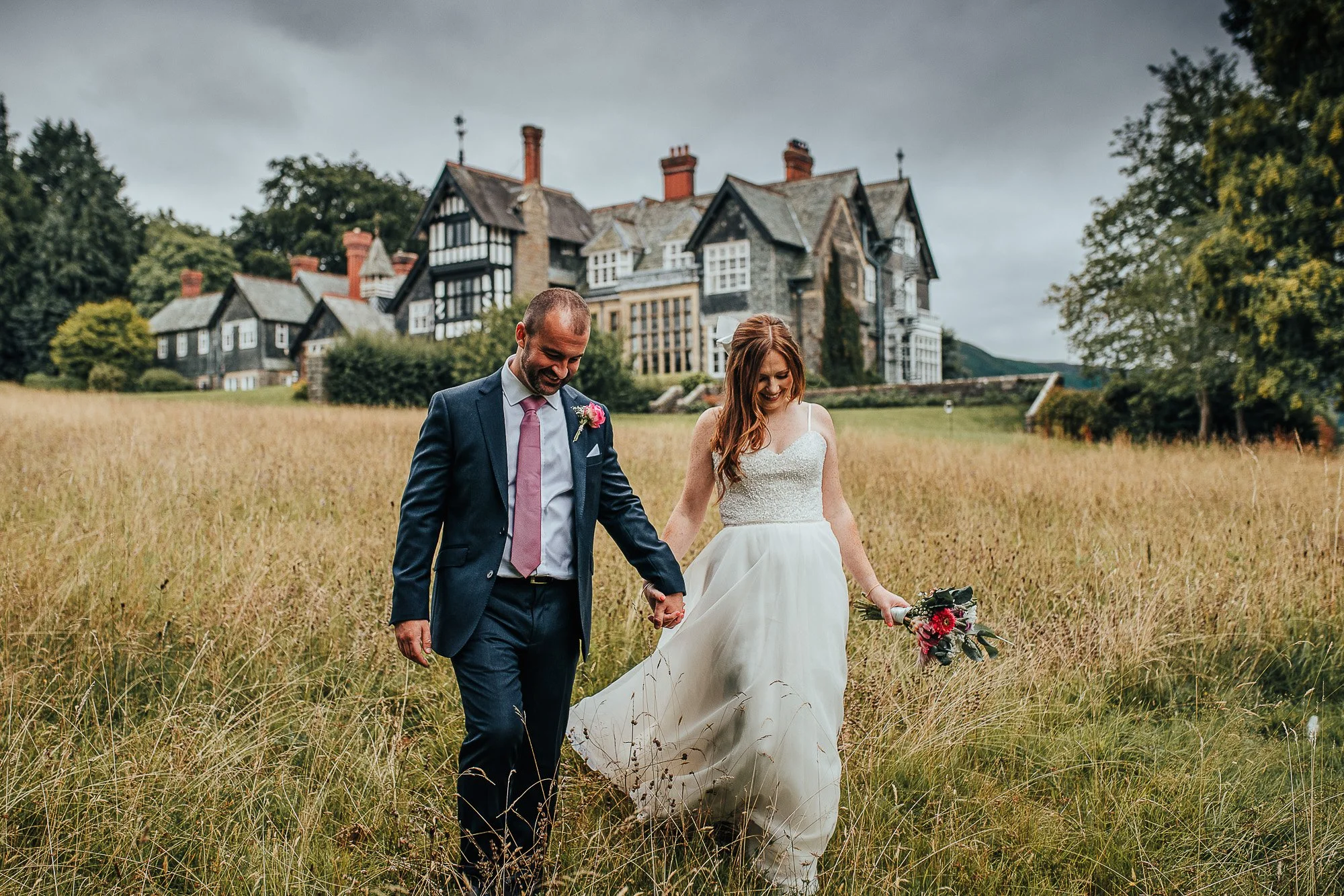 Bride and groom walking hand in hand through a grassy field with a manor house in the background, overcast sky.