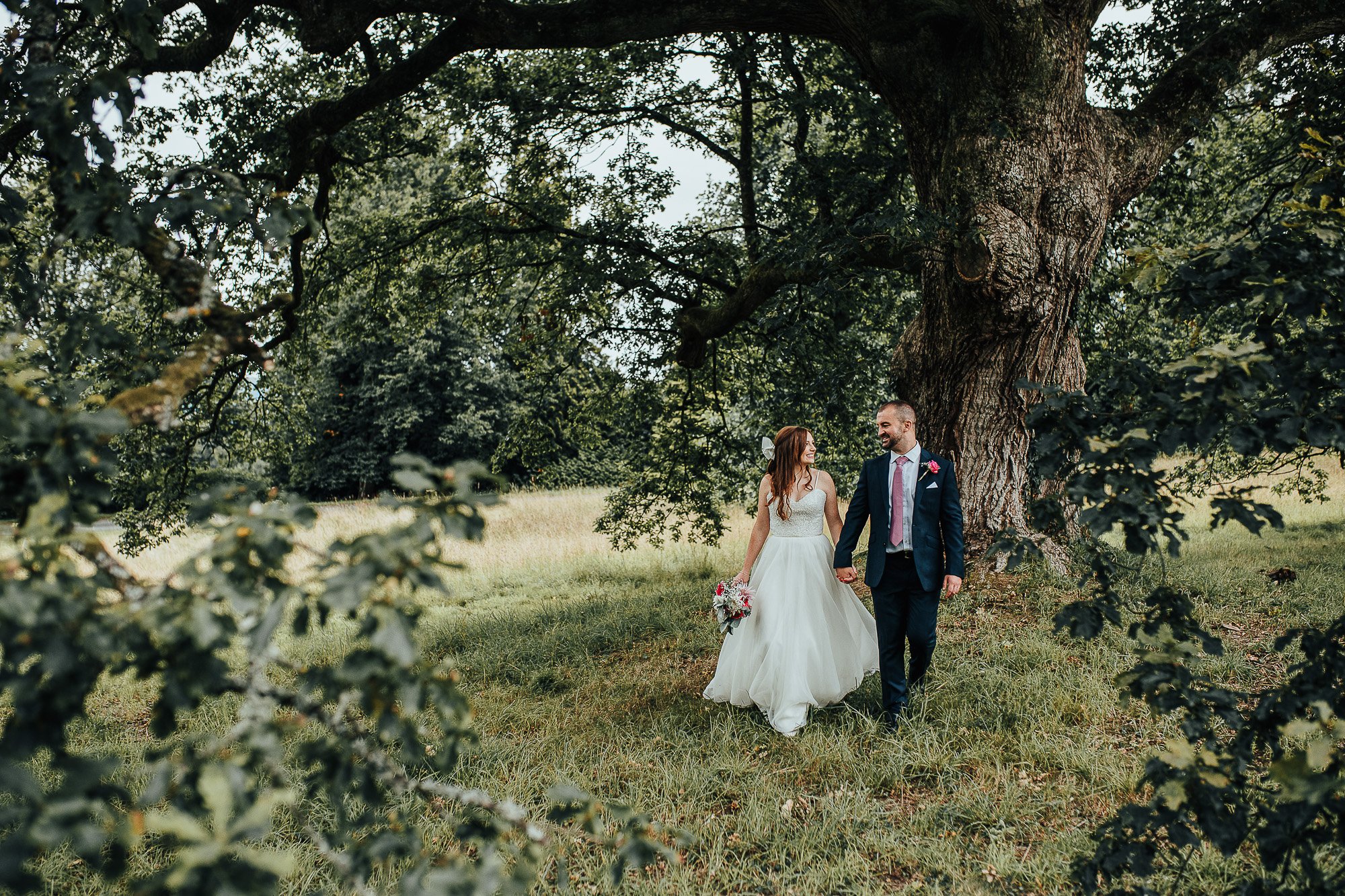 Bride and groom walking outdoors under a large tree, surrounded by greenery and grass, bride holding a bouquet.