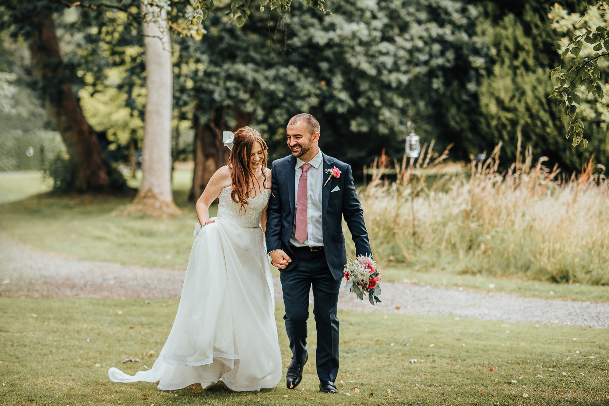 Bride and groom walking on grass, hand in hand, with the bride holding her gown and the groom holding a bouquet, in a green outdoor setting.