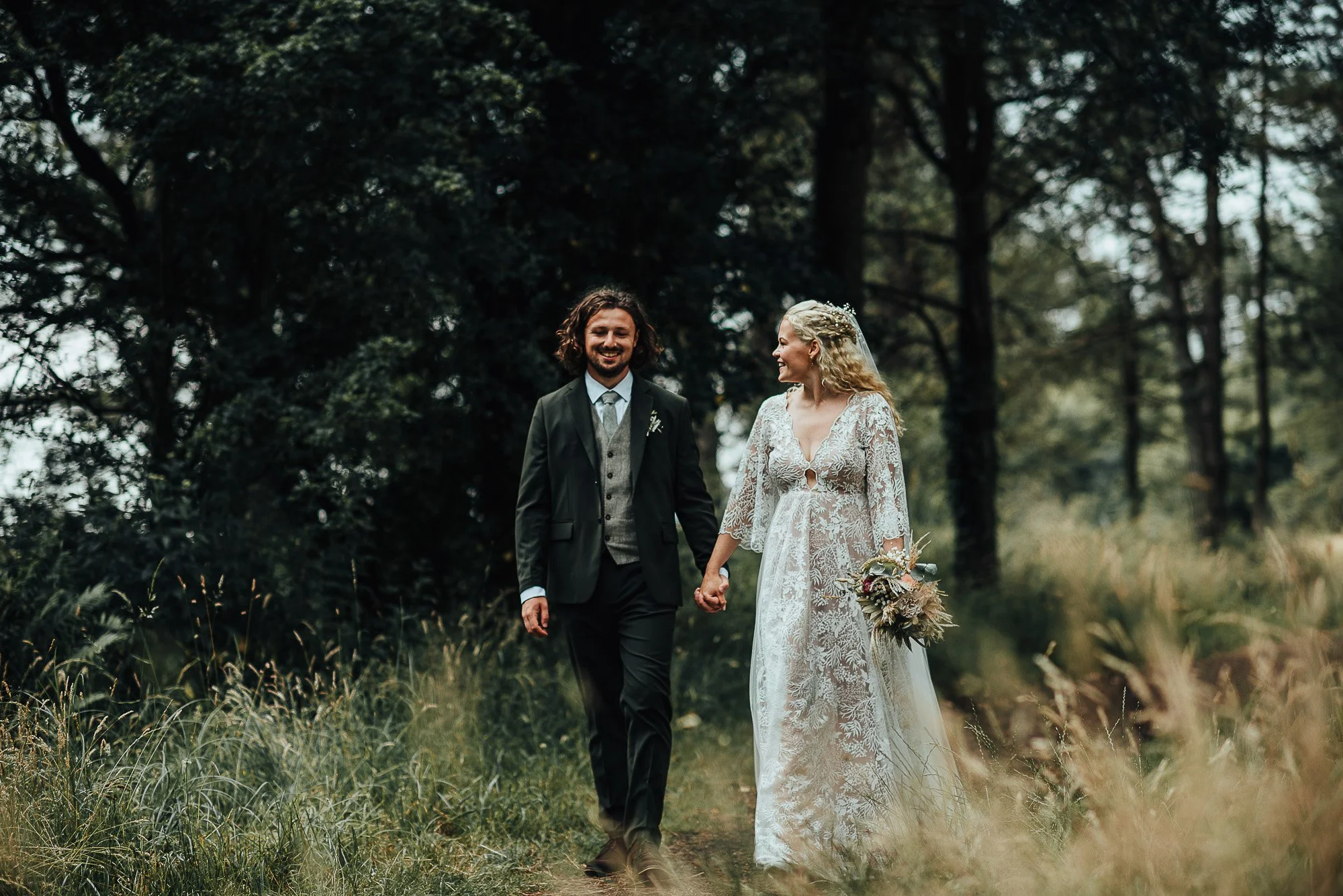 A bride and groom walking hand in hand through a forest, smiling at each other. The bride is wearing a lace wedding dress and holding a bouquet, while the groom is in a suit.