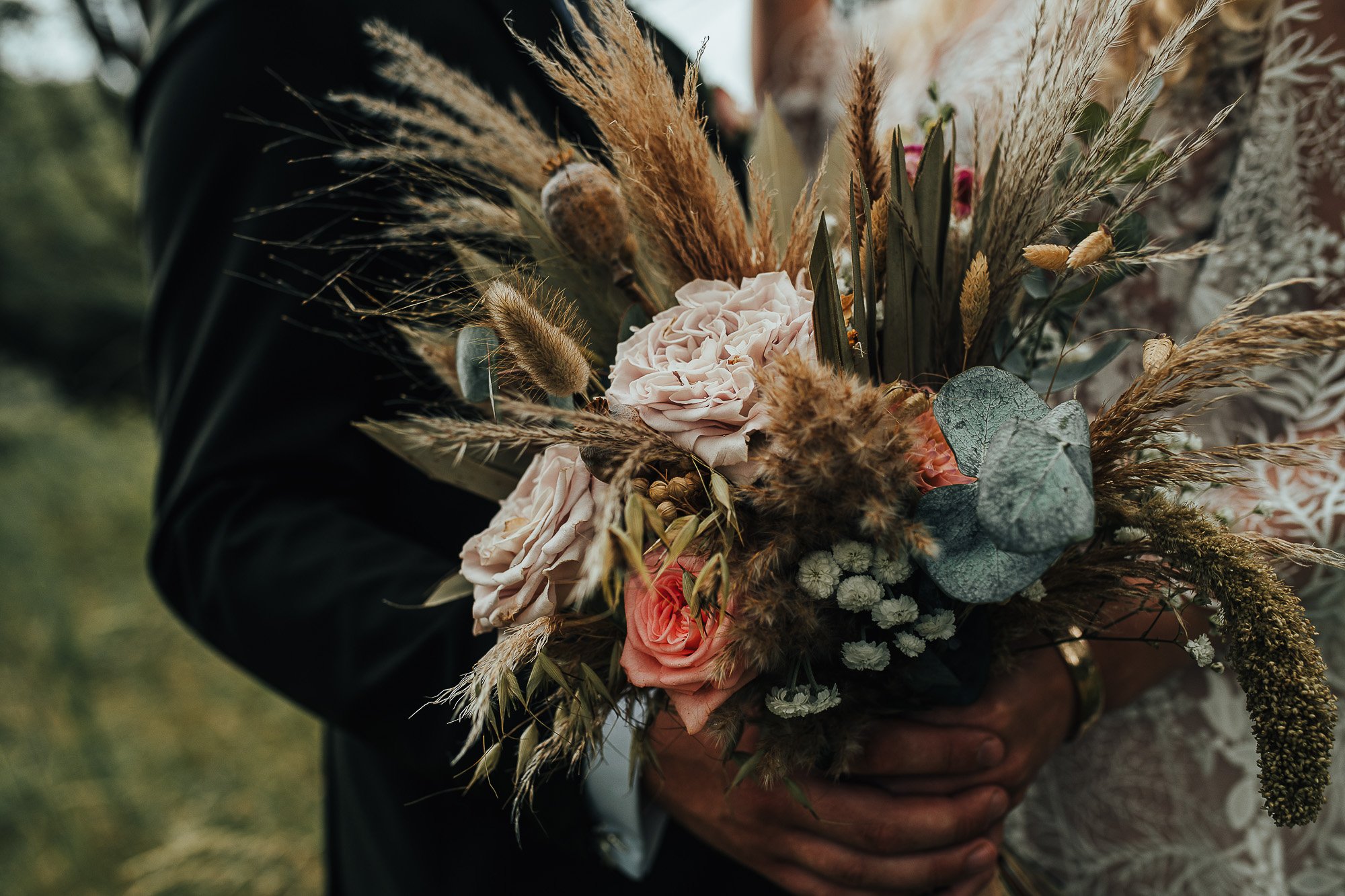 Close-up of a bride and groom holding a rustic bouquet with pink roses, dried grasses, and greenery.