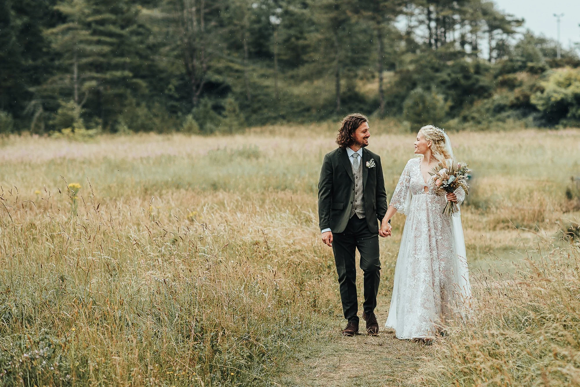 Bride and groom walking in a grassy field, dressed in wedding attire, holding hands and smiling, surrounded by trees.
