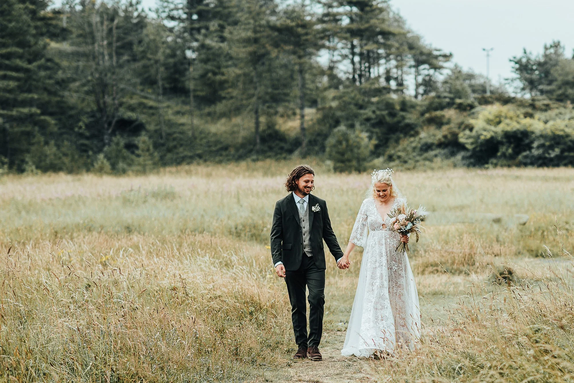 A couple walks hand in hand through a grassy field, with the woman in a white lace dress holding a bouquet and the man in a dark suit, against a backdrop of trees.
