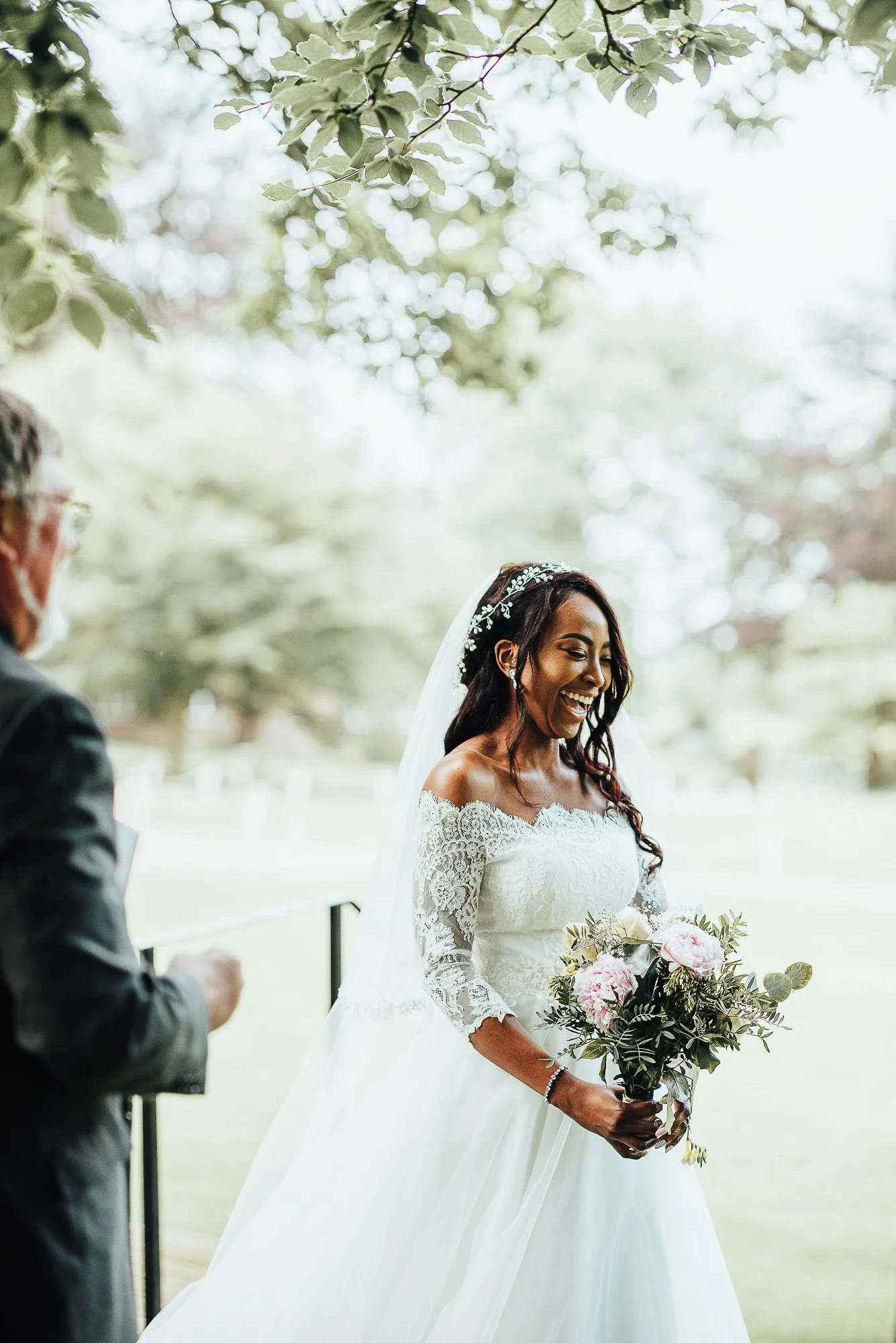 Bride in white lace wedding dress holding bouquet under tree outdoors, smiling at a man in a suit.