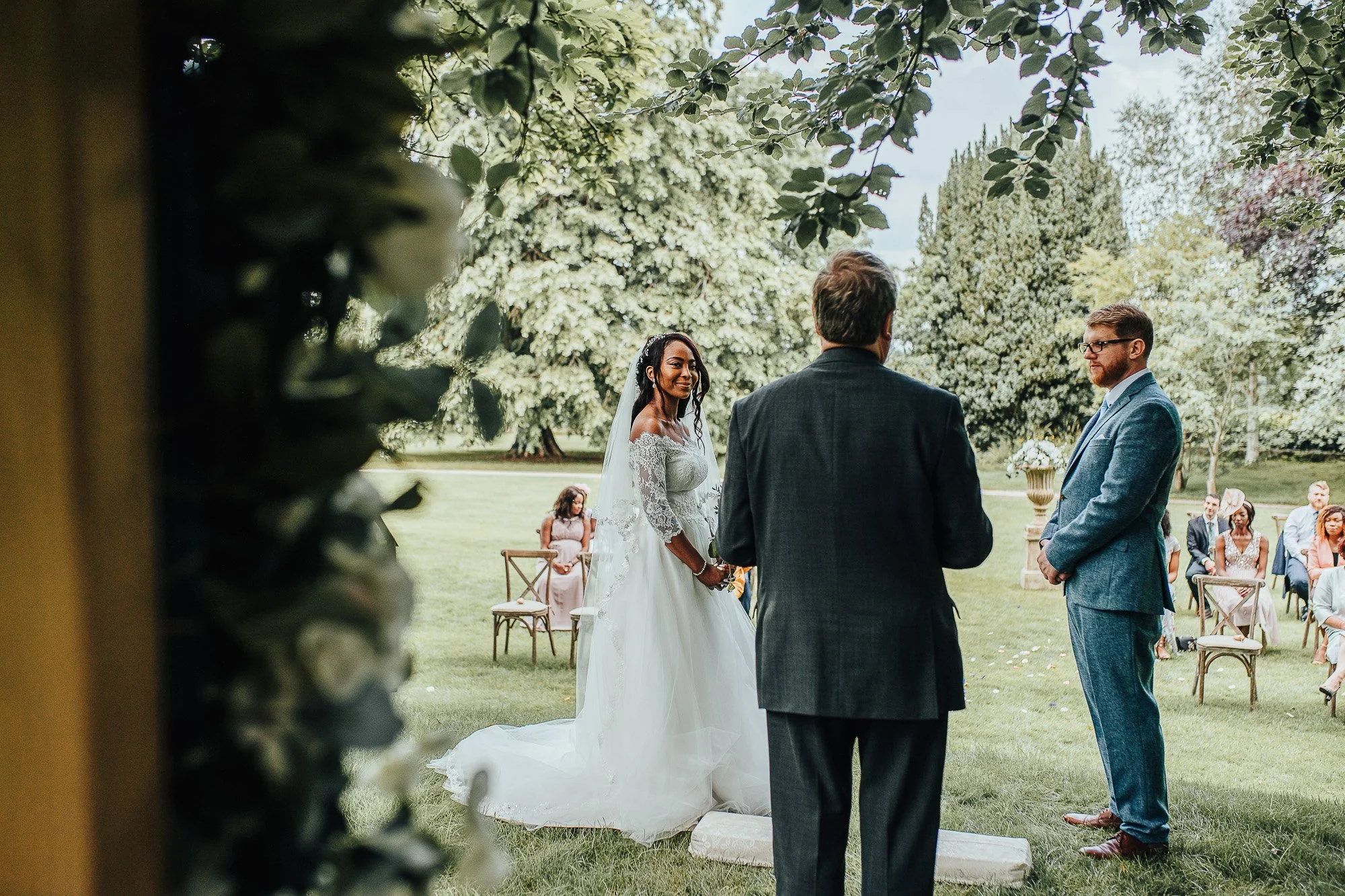 An outdoor wedding ceremony with bride in a white dress facing a groom in a blue suit, officiant standing between them, guests seated in the background on a grassy area with trees.