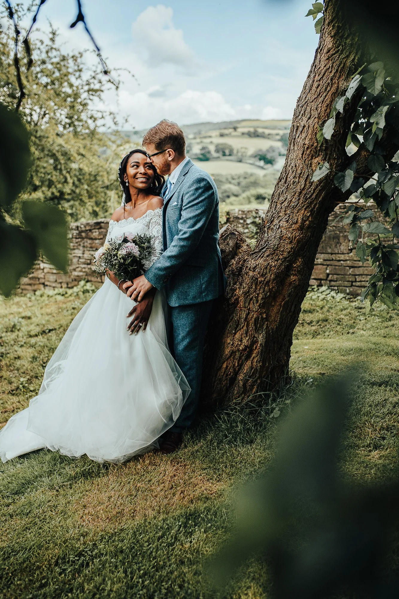 Bride and groom pose outdoors by a tree, with scenic landscape background. Bride in white dress holding a bouquet, groom in blue suit.