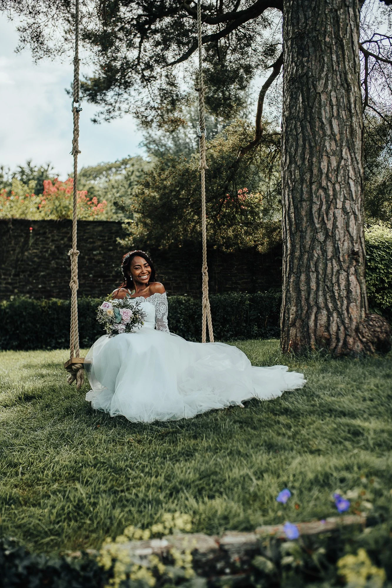 Bride in white gown sitting on a swing in a garden, holding a bouquet.