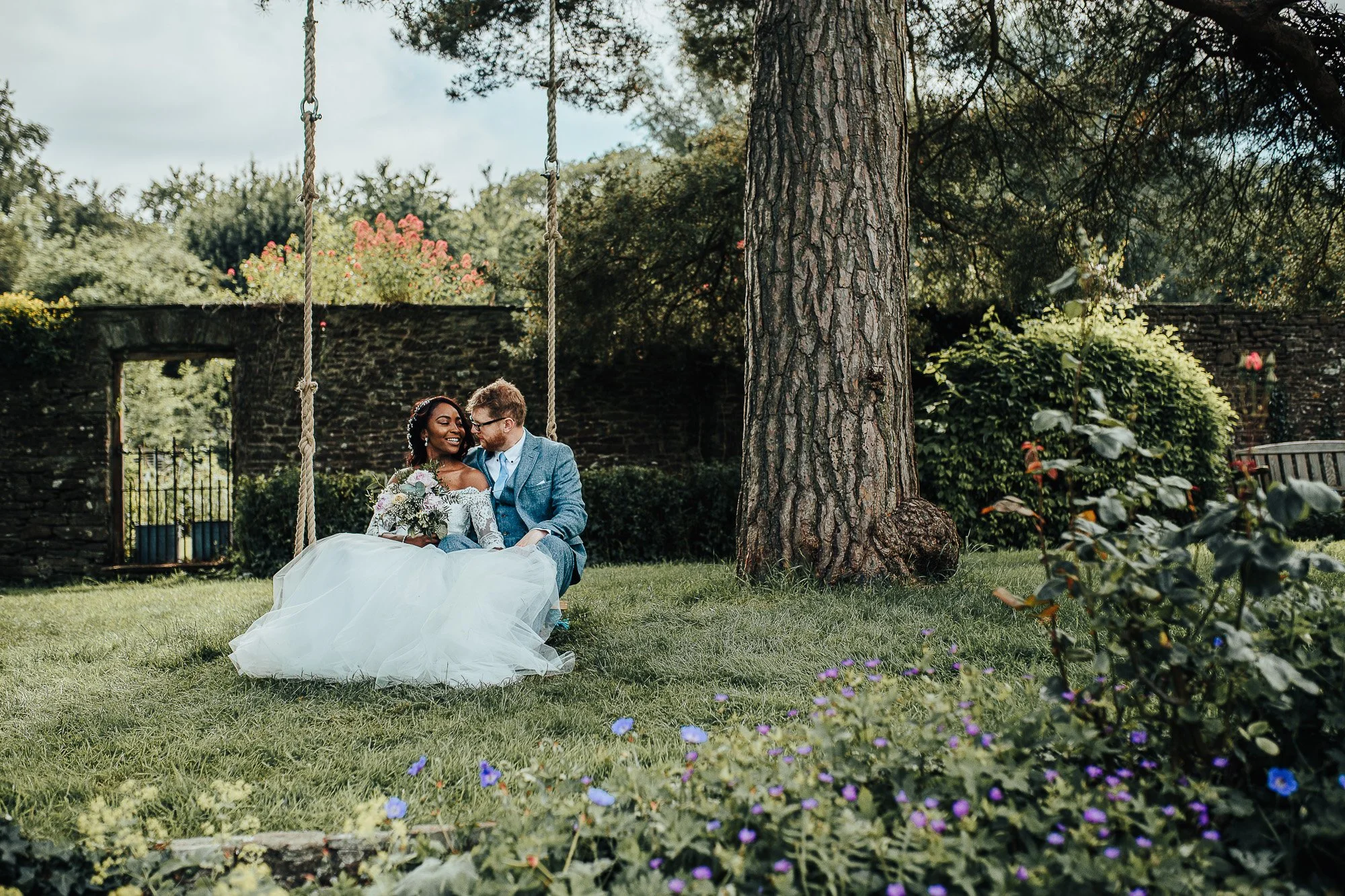 Bride and groom sitting on a swing under a tree in a garden, surrounded by greenery and flowers.
