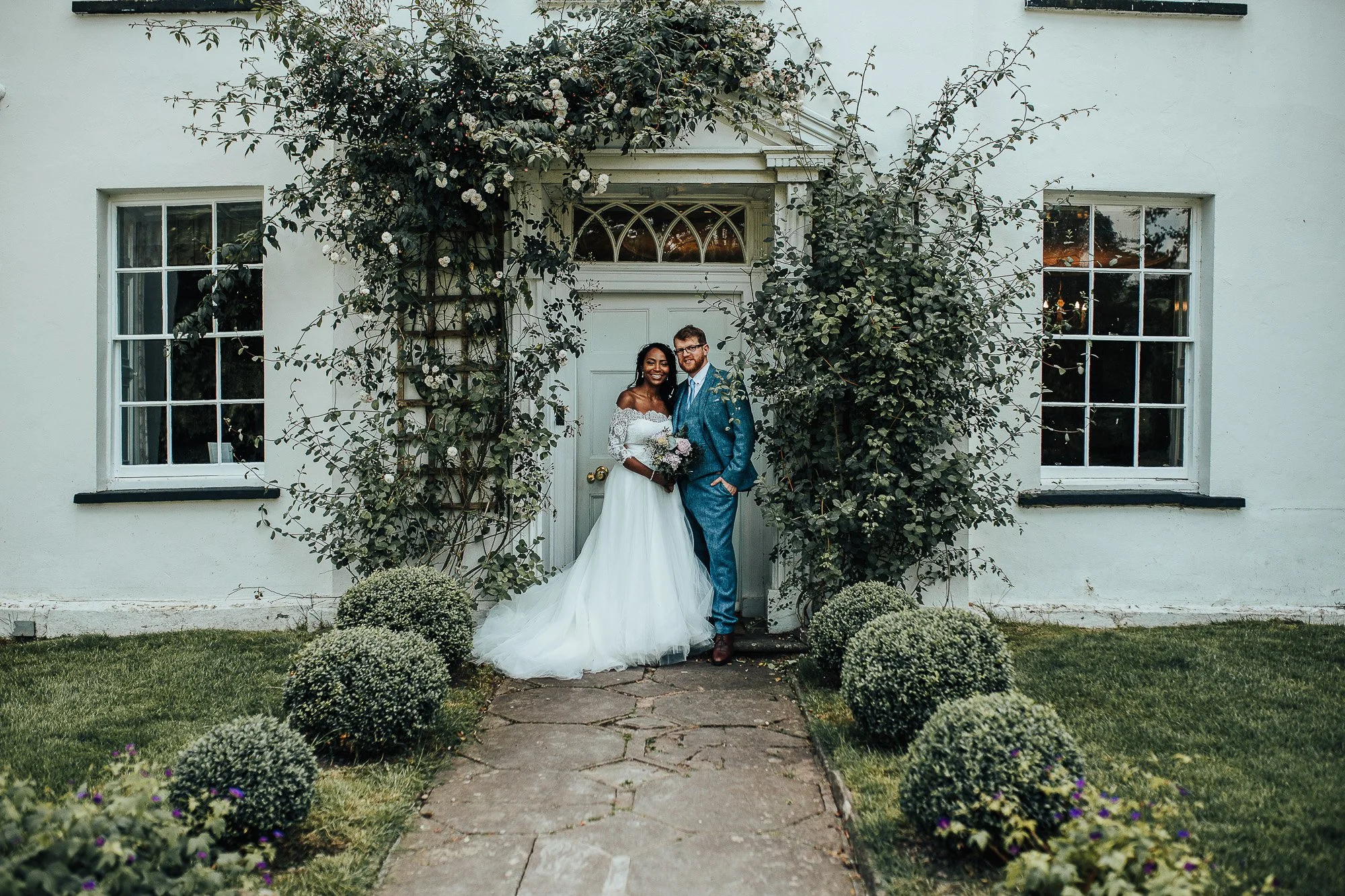 Bride and groom standing in front of a white house with a green garden, posing under a vine-covered archway.