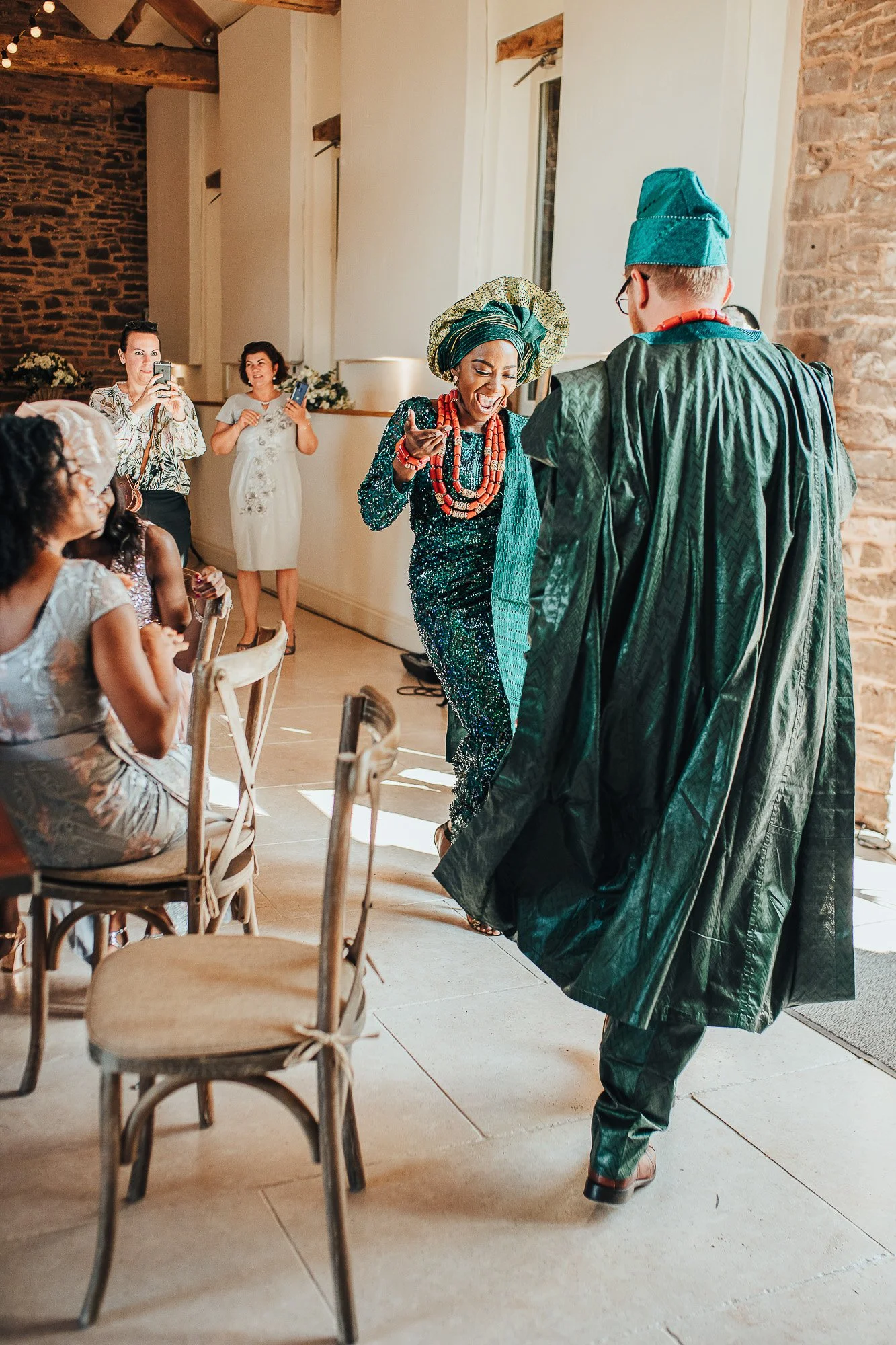A couple wearing traditional green attire dances at a cultural event, surrounded by seated guests and photographers.