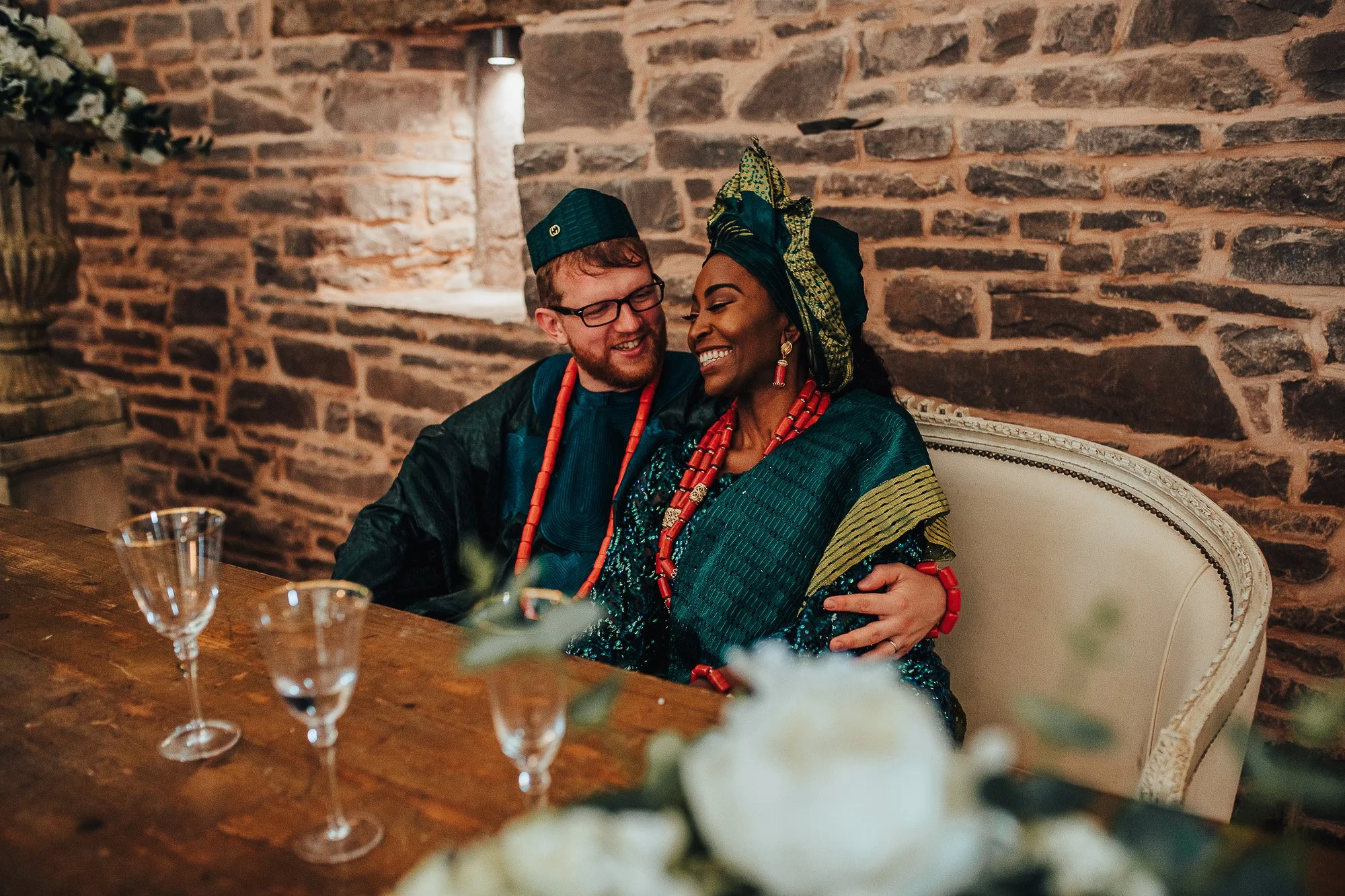 Couple wearing traditional attire seated and smiling, on a rustic stone background, with glasses on the table.