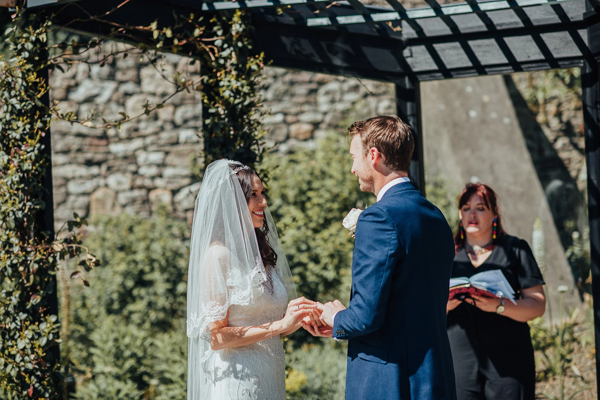 A bride and groom exchanging vows outdoors under a pergola, with an officiant standing behind them. The bride is wearing a white dress and veil, and the groom is in a blue suit. The scene is set in a garden with greenery and a stone wall in the backg