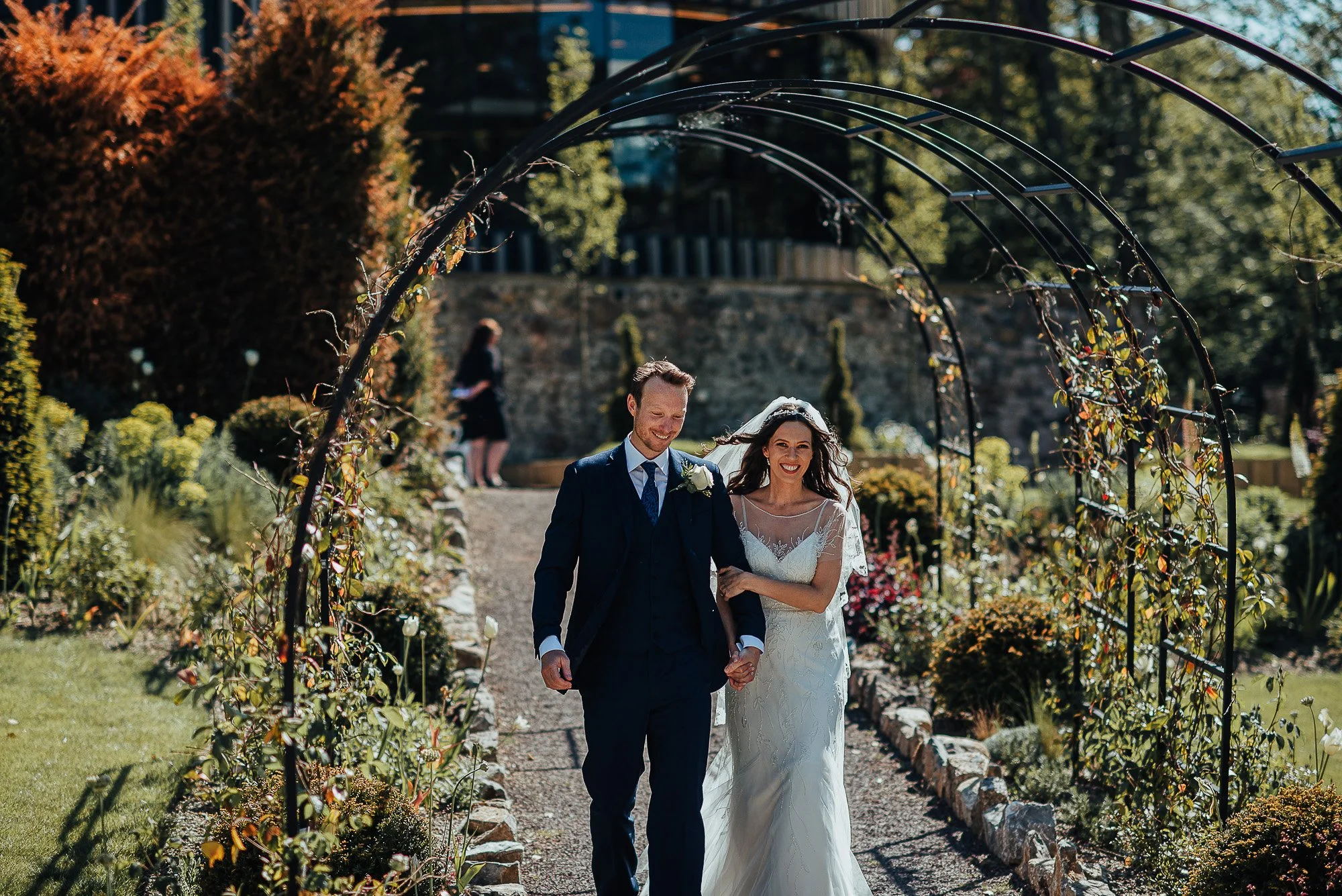 Bride and groom walking through a garden archway, smiling, with greenery and flowers around.