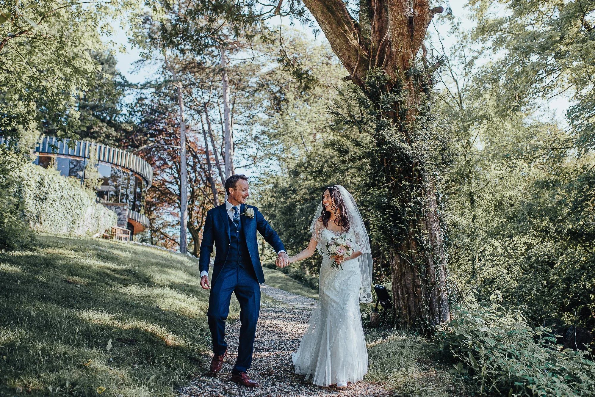 Bride and groom walking hand in hand down a tree-lined path, with the bride wearing a white wedding dress and holding a bouquet, and the groom in a blue suit. They are smiling at each other. A building and green foliage are visible in the background.