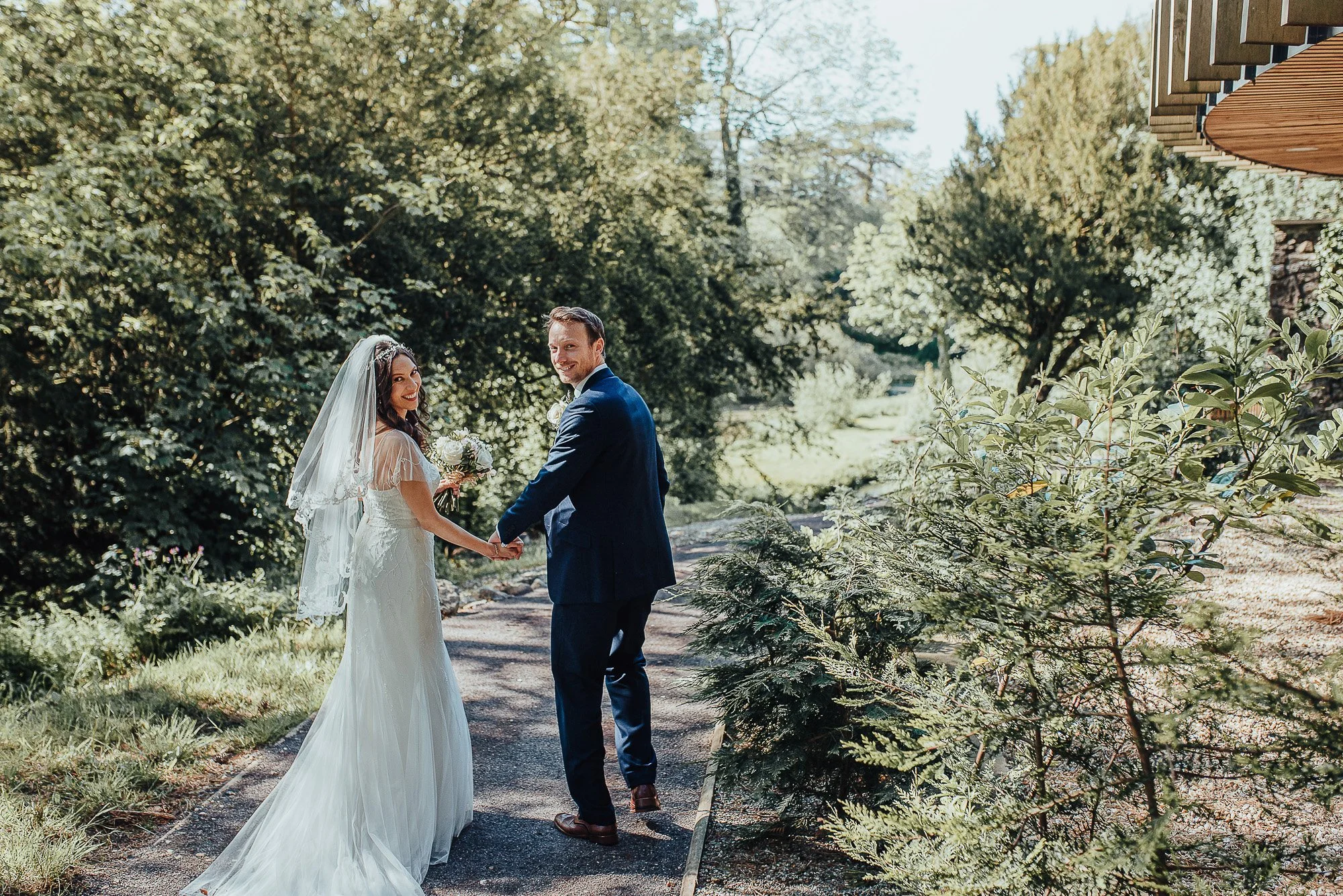 Bride and groom holding hands on a garden path, surrounded by greenery, with the bride wearing a white dress and veil and the groom in a dark suit.