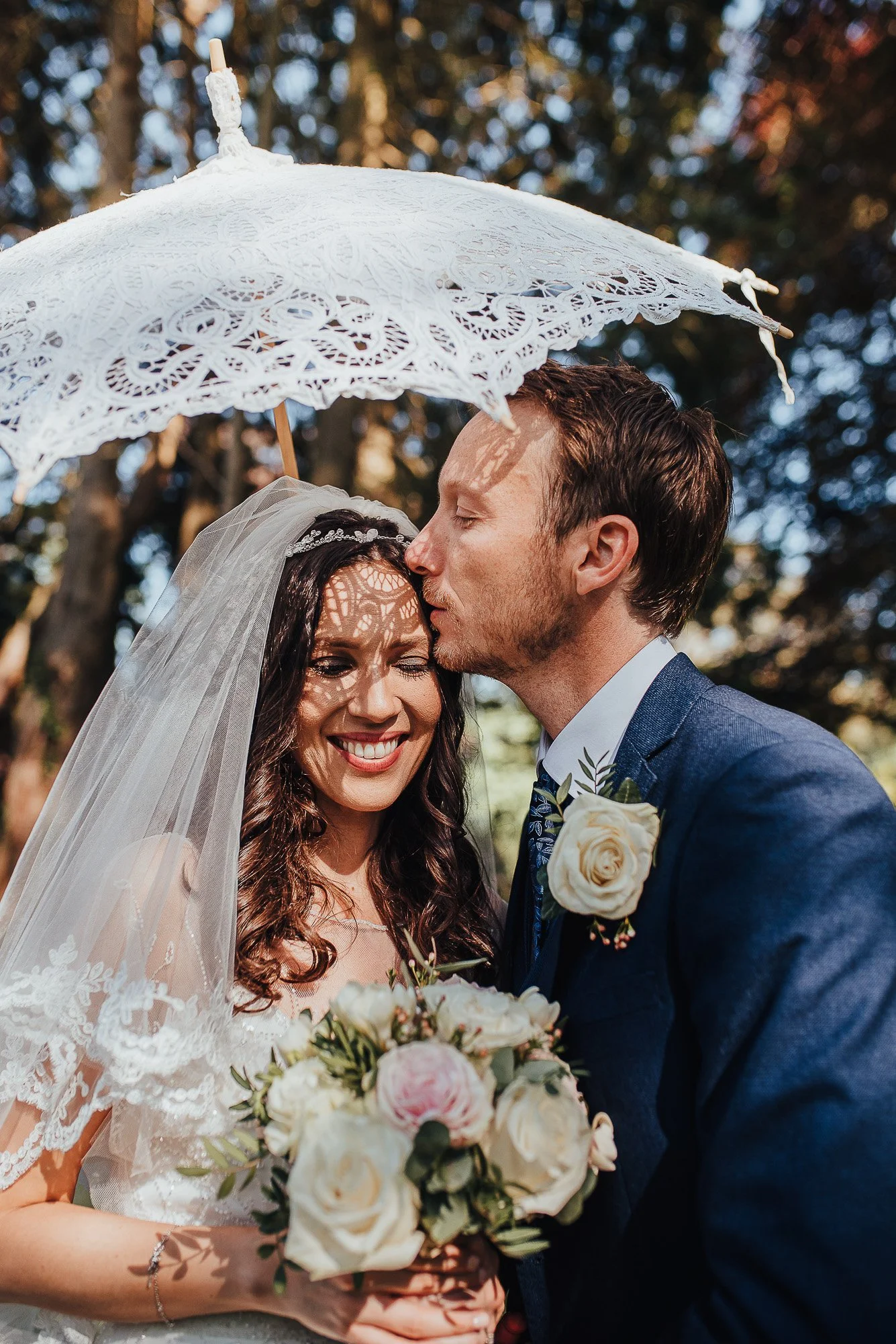 Bride and groom under a lace umbrella, bride holding a bouquet, groom kissing her forehead.
