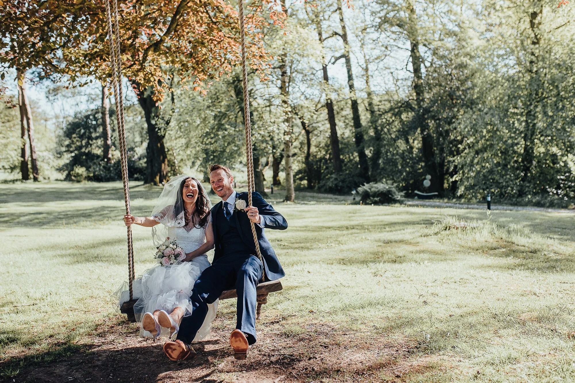 Bride and groom sitting on a swing in a garden, both smiling with joy. The bride is in a white gown holding flowers, and the groom is in a navy suit. The background features trees and greenery, suggesting a peaceful, outdoor setting.