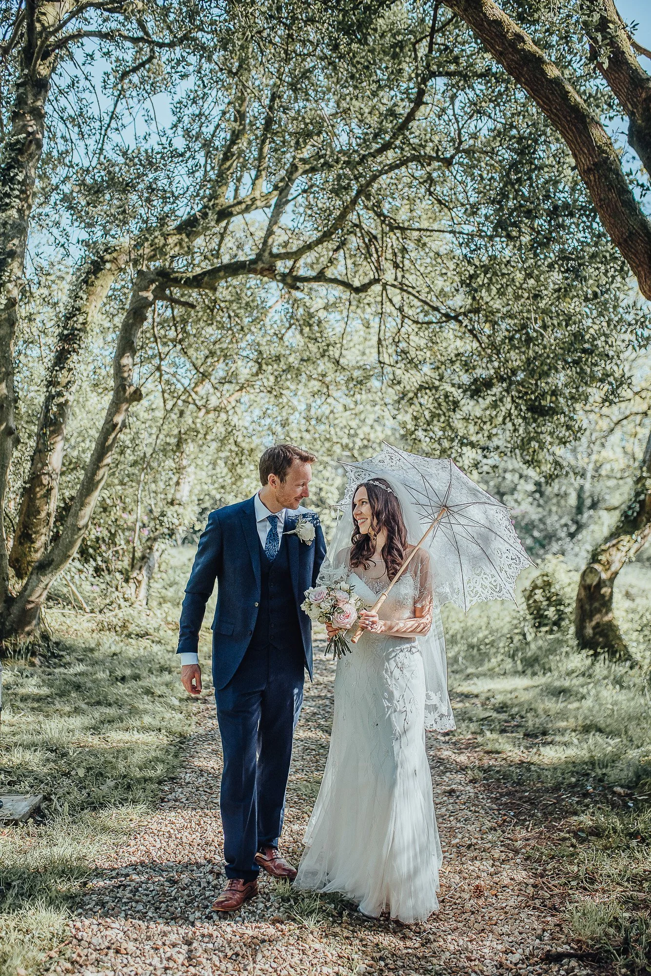 Bride and groom walking under trees, bride holding a parasol.