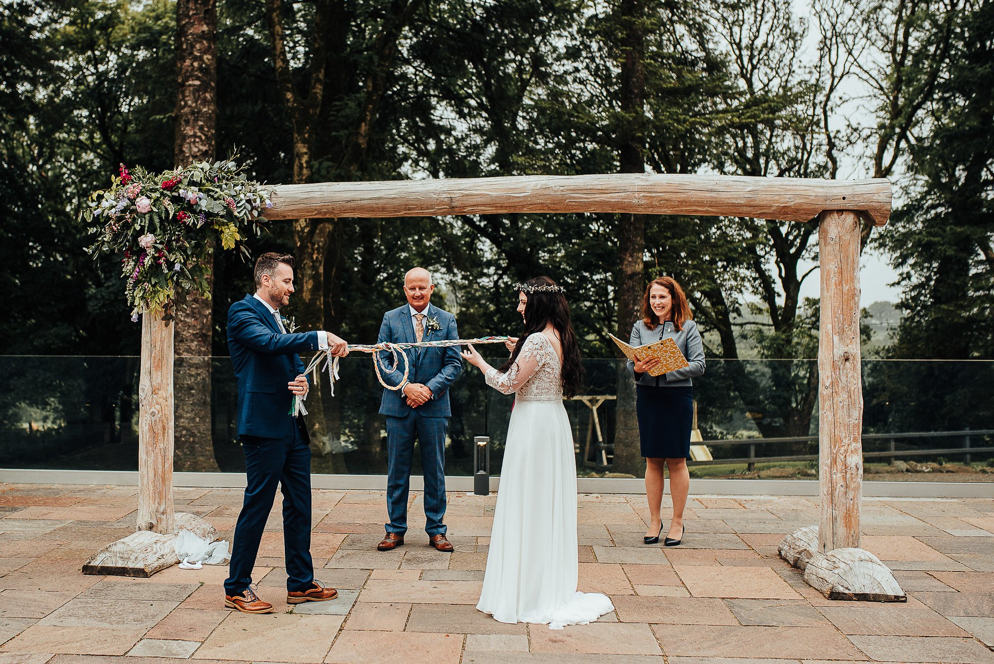Wedding ceremony with couple holding ropes, officiant, and witness under wooden arch adorned with flowers, outdoors.