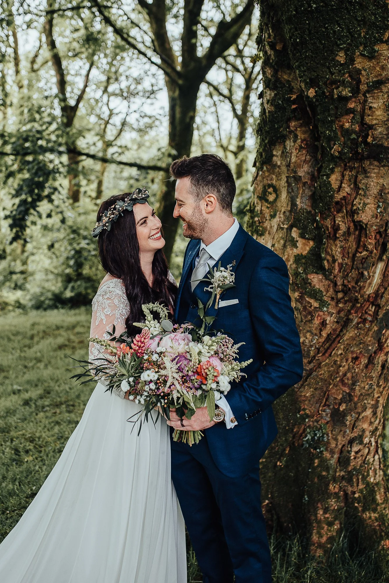 Bride and groom smiling at each other in a forest setting, bride holding a large bouquet of flowers, groom in a dark suit.