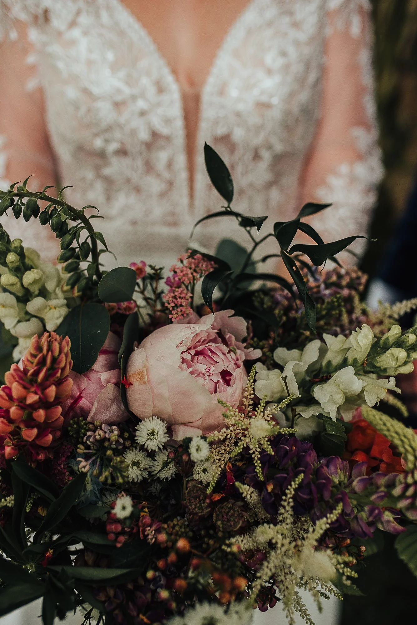 Close-up view of a bride's bouquet with pink peonies, white and purple flowers, and green leaves, held by a person in a lace dress.