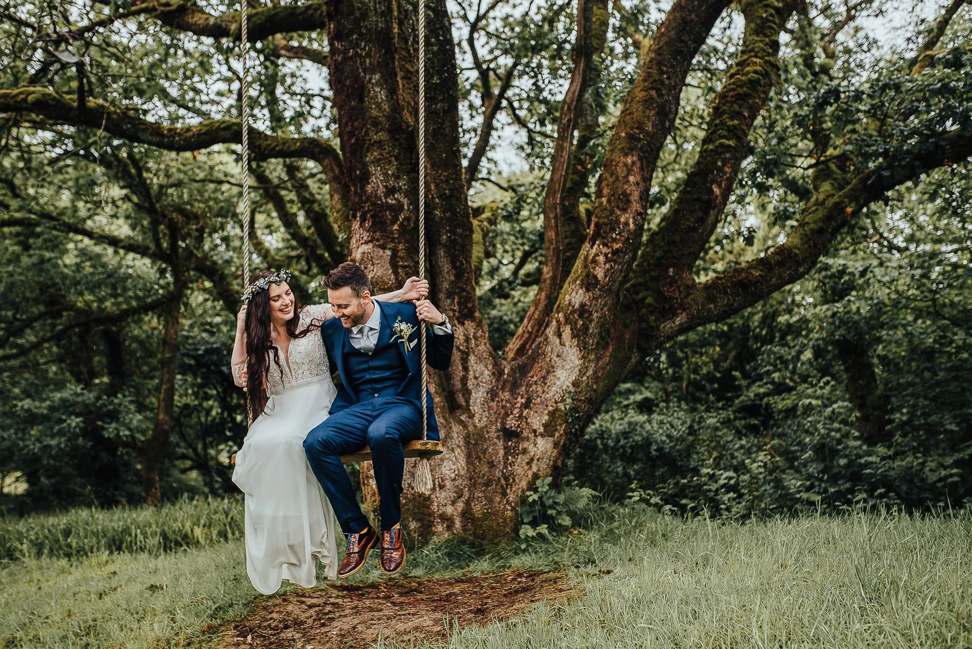A newlywed couple sitting on a swing in a wooded area, with the bride wearing a white dress and floral crown, and the groom in a suit, in a joyful and romantic pose.