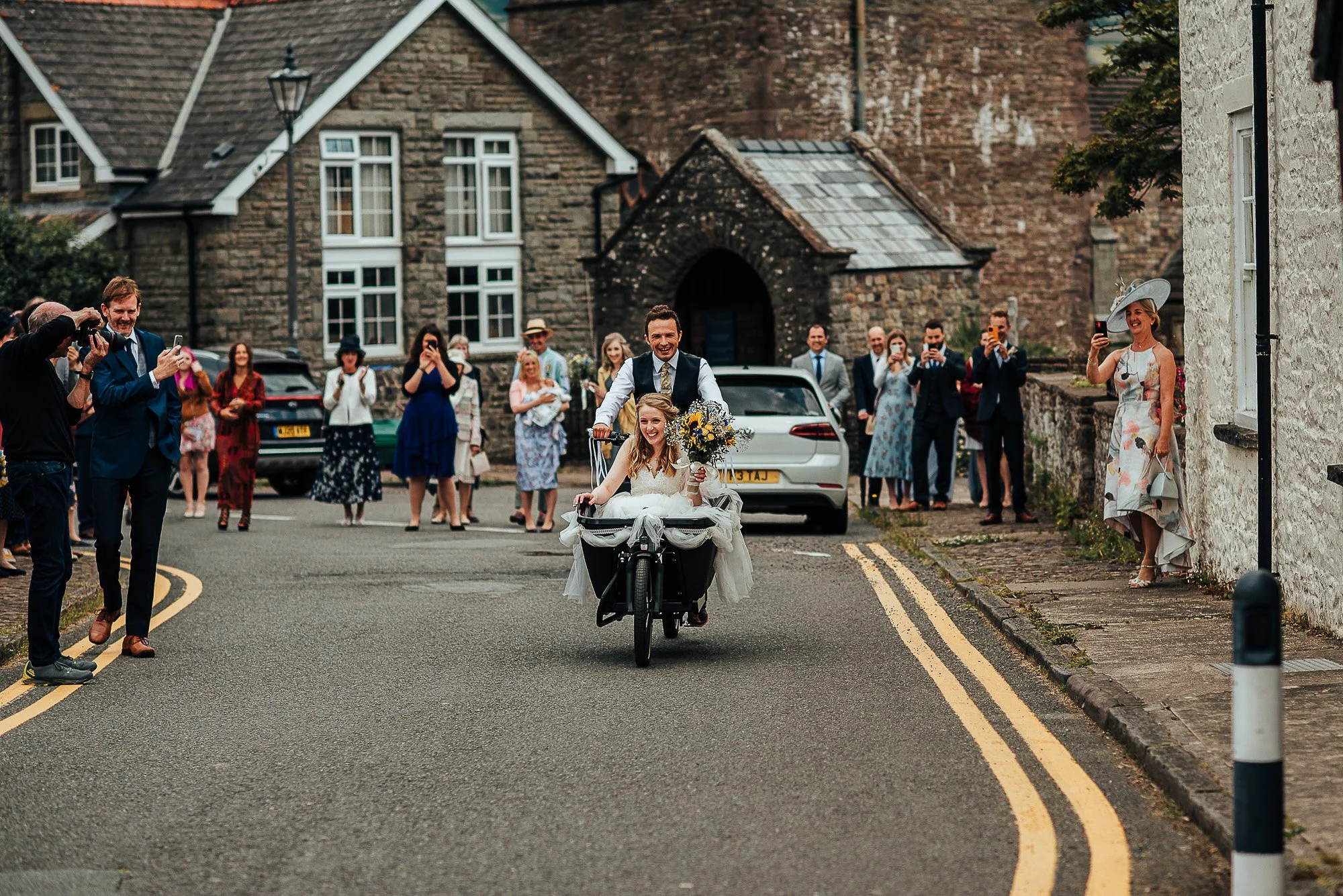A bride in a white dress sitting in a cargo bike basket, holding a bouquet, while a groom, dressed in a suit, pedals. They are on a street with a gathering of people taking photos and cheering. Stone buildings and a parked car are visible in the back
