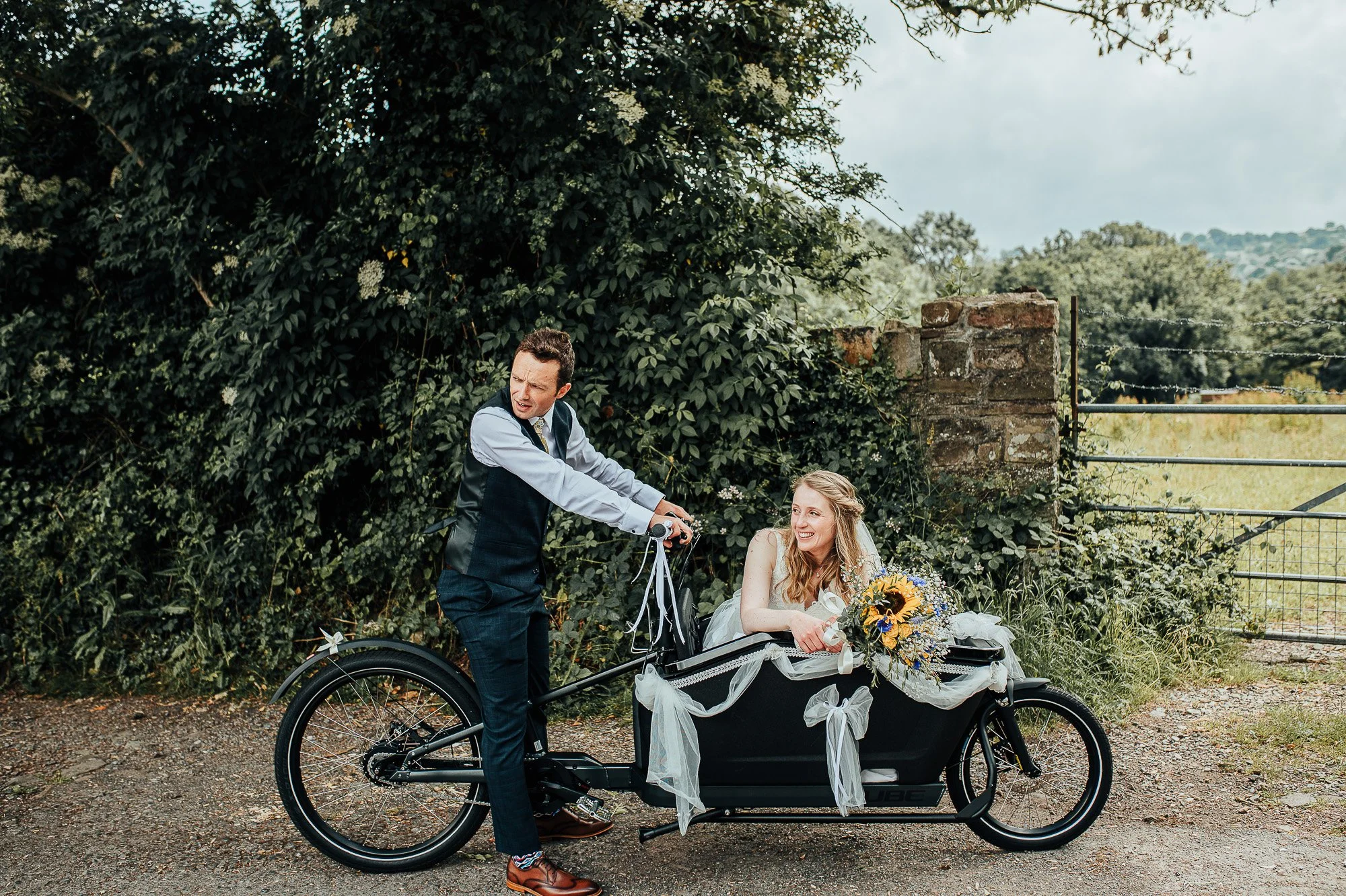 A bride sitting in a decorated cargo bike with a bouquet of sunflowers, while a man stands next to her, mimicking cycling; scenic outdoor background.